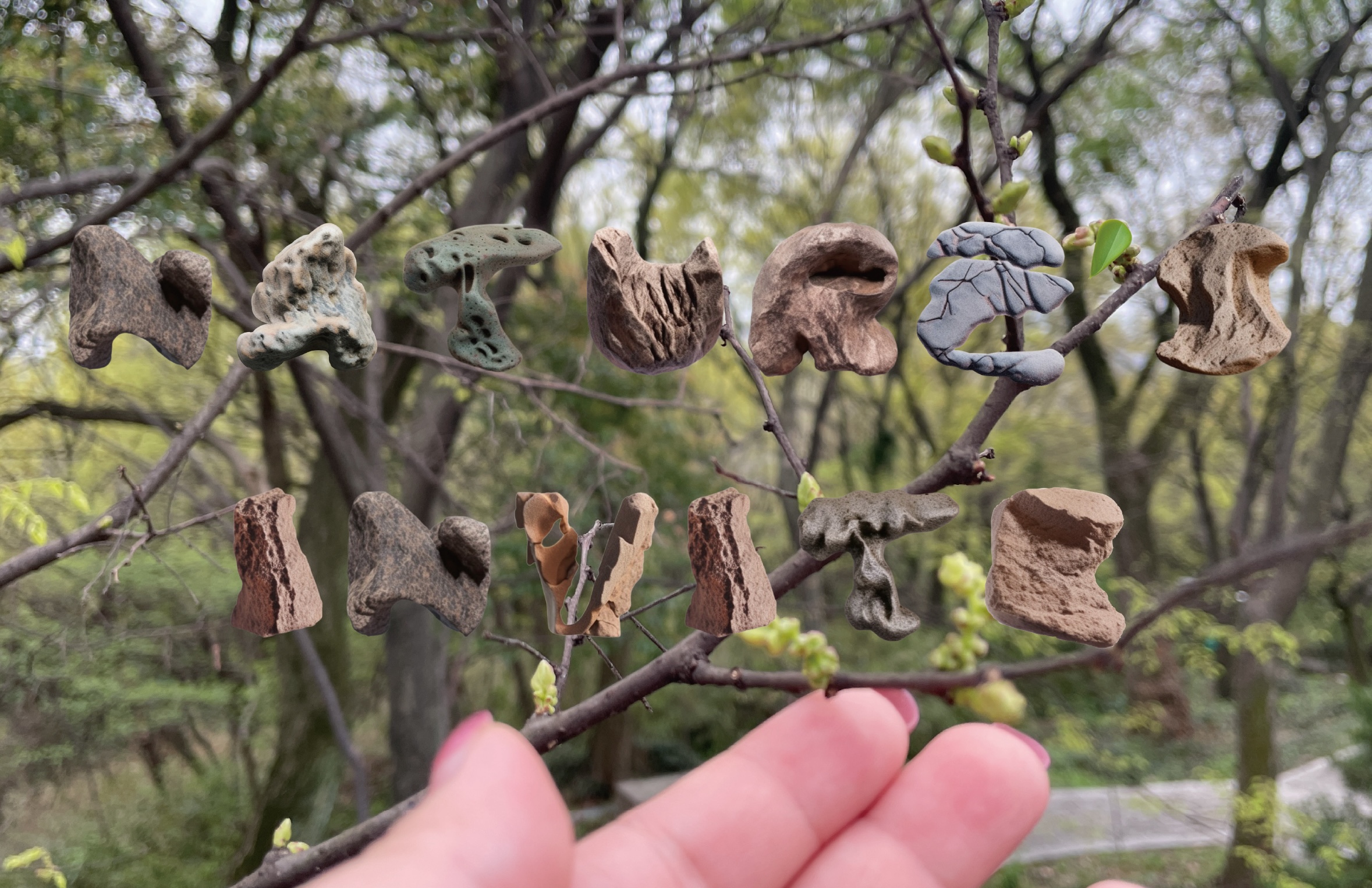 A hand holds a branch in a forest setting, with organic-shaped clay or stone pieces suspended on the twigs to form the words 'Nature Nurture' against a blurred background of trees.