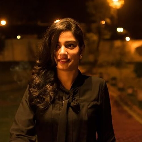 A woman with long dark hair stands outdoors at night, illuminated by warm lighting. She is wearing a black blouse with a bow detail and looks slightly to the side with a smile.