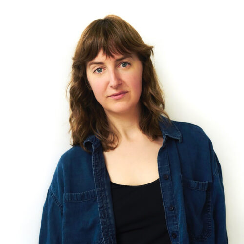 A woman with wavy brown hair wearing a dark blue denim shirt over a black top poses against a plain white background. She is looking directly at the camera with a calm, neutral expression.