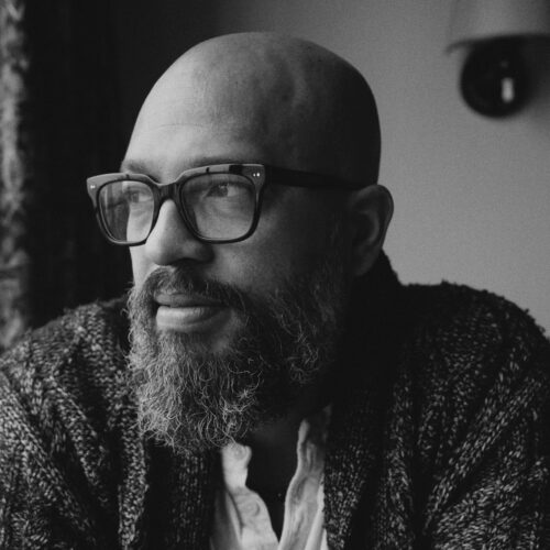 A close-up black and white portrait of a bald man with a thick beard, wearing oversized glasses. He appears to be gazing thoughtfully out of a window, with soft lighting highlighting his facial features. He is dressed in a textured, dark cardigan over a light-colored shirt. The background features a blurred, patterned curtain.