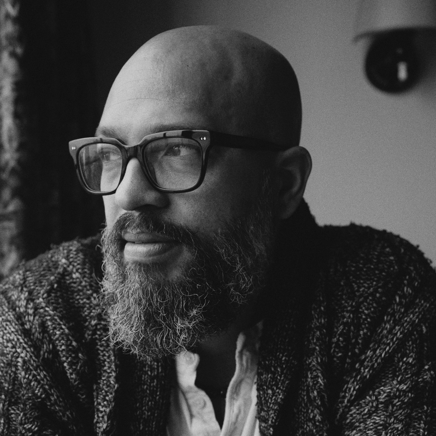 A close-up black and white portrait of a bald man with a thick beard, wearing oversized glasses. He appears to be gazing thoughtfully out of a window, with soft lighting highlighting his facial features. He is dressed in a textured, dark cardigan over a light-colored shirt. The background features a blurred, patterned curtain.