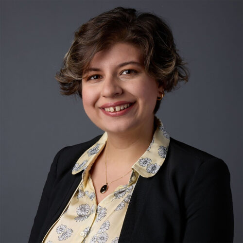 A woman with short, wavy dark-blond hair smiles against a dark gray backdrop. She wears a black blazer over a pale yellow floral blouse with a necklace.