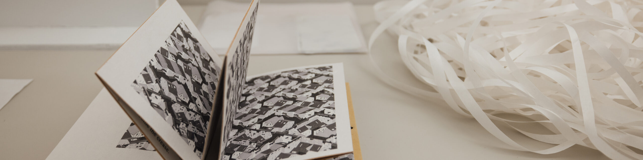 An open book with black and white illustrations of houses sits on a table. The pages are folded in a way that creates a 3D effect. Nearby, there is a bundle of white strips of paper, possibly for binding or crafting. A couple of plain sheets are also visible in the background.
