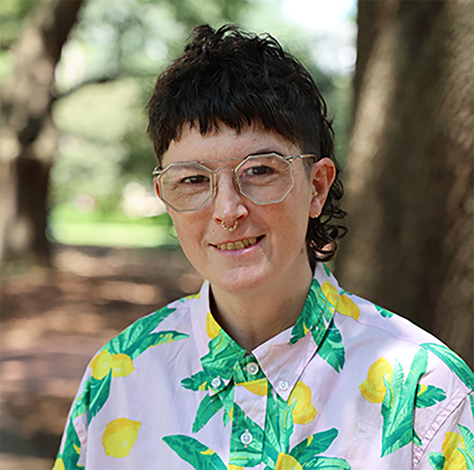 A person with short dark hair, glasses, and a septum piercing smiles while standing outdoors among trees. They are wearing a light pink shirt patterned with large yellow lemons and green leaves.