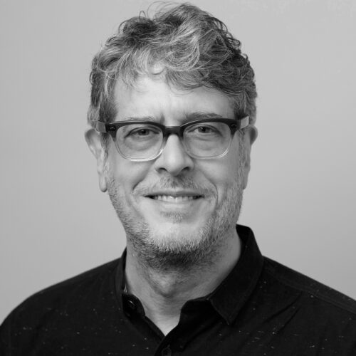 A smiling man with curly gray hair and glasses is facing the camera. He is wearing a dark shirt and appears friendly, set against a plain light background.
