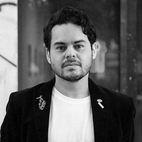 A man with dark, curly hair and a beard is standing in front of a blurred background. He is wearing a black jacket over a white t-shirt, with a couple of pins on his lapel. The image is in black and white, capturing a serious expression.
