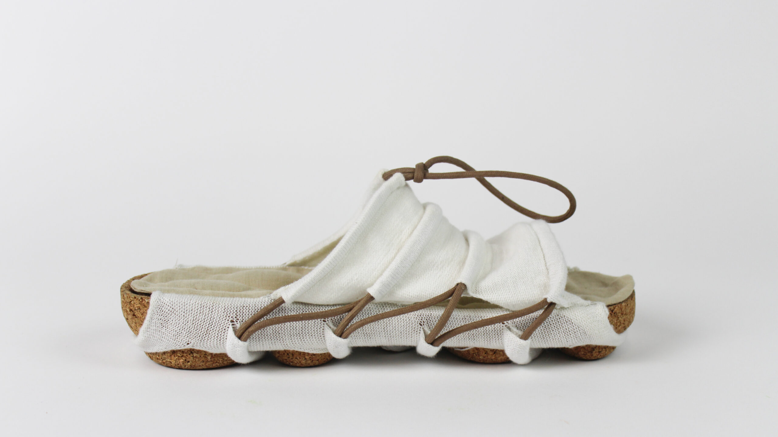 Side view of a finished handcrafted sandal with a cork sole, white textile body, and brown cord lacing, photographed against a plain white background.