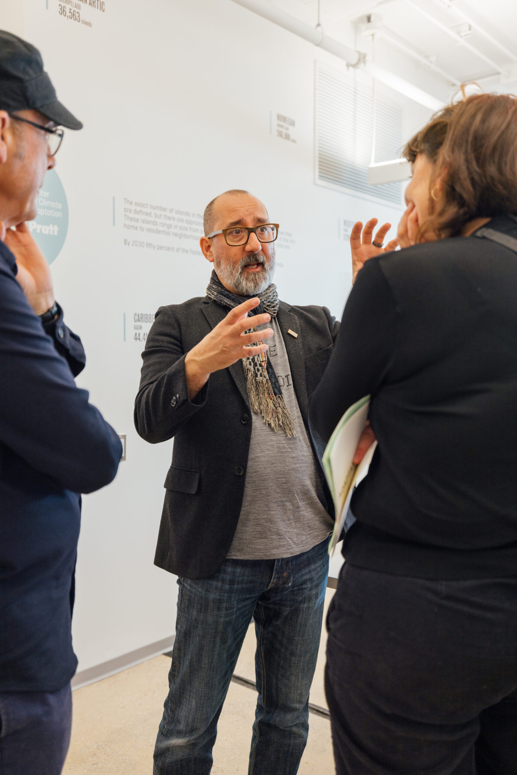 A man with glasses and a beard is animatedly speaking to two individuals in a casual setting. He wears a blazer over a t-shirt and a scarf. One person is partially visible on the left, while the other, also partially visible, is listening intently, holding a folder or notebook. In the background, a wall displays text and information, suggesting an exhibition or educational environment.