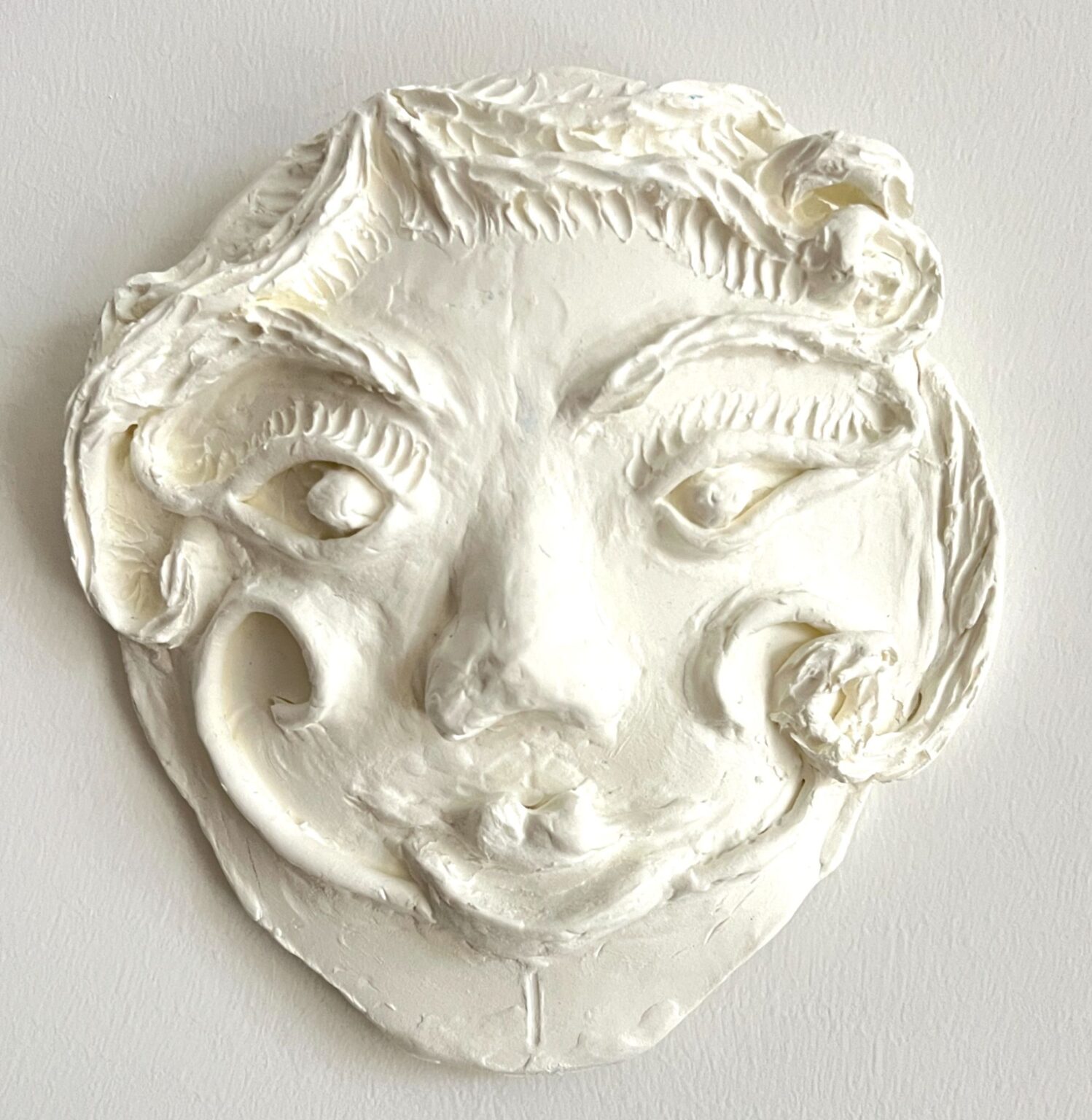 A white sculpted face mask with stylized features, including large eyes, curled eyebrows, a pronounced nose, and swirling decorative shapes along the cheeks and hairline. The surface has a textured, hand-molded appearance.