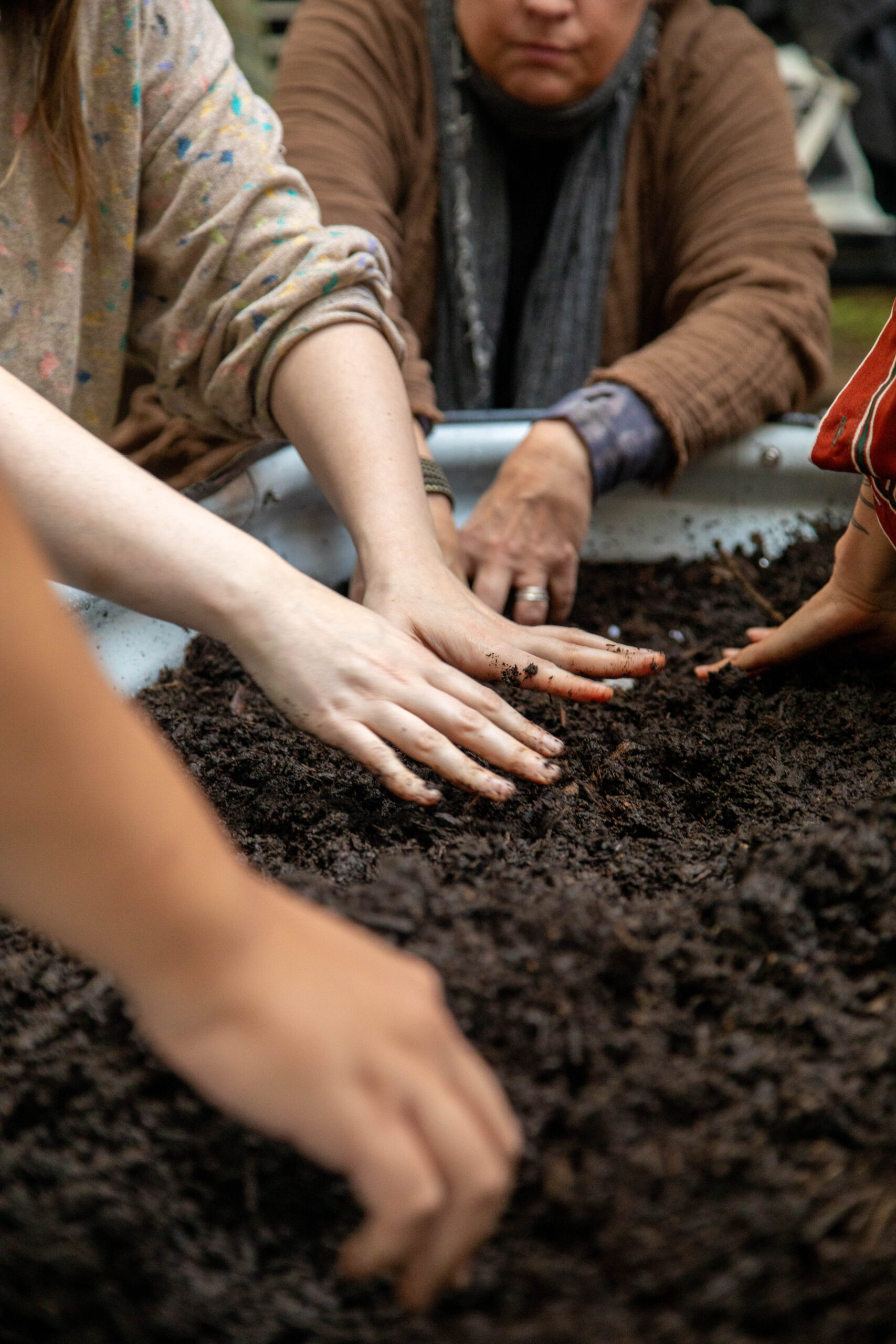 Several hands are reaching into a large container filled with dark, rich soil. The foreground shows a close-up of the hands, some with dirt on them, actively mixing or feeling the soil. In the background, a few individuals are partially visible, focused on the activity, with a blurred focus on their upper bodies and faces. The setting appears to be outdoors or in a casual group environment, suggesting a community gardening or planting activity.