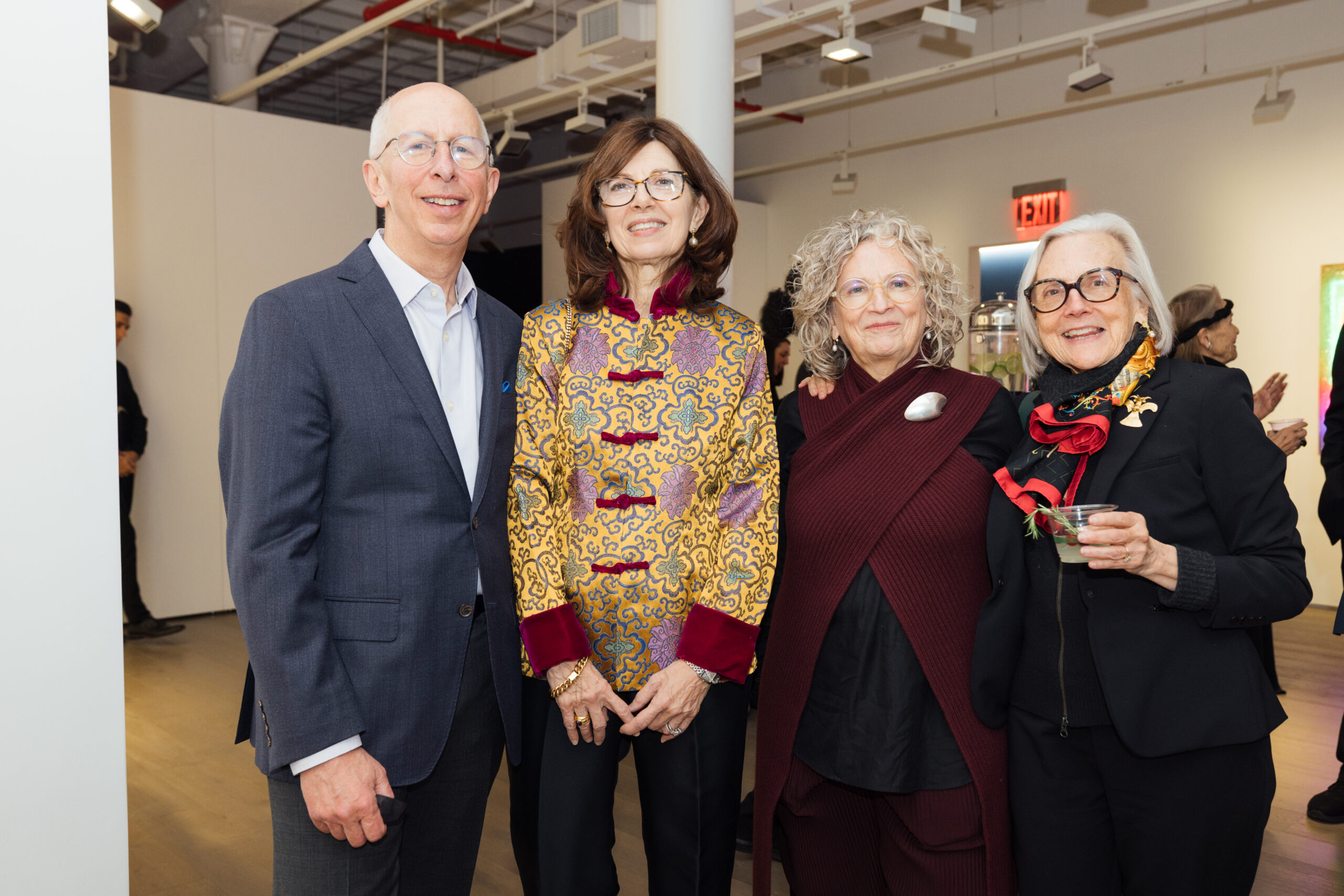 A group of four adults stands together, smiling for the camera in an art gallery setting. The man on the left wears a blue blazer over a white shirt. The second person, a woman, is dressed in a colorful patterned top with red accents. The third person has curly hair and wears a maroon outfit, while the fourth woman is in black attire and holds a drink, accessorized with a colorful scarf. Background features art pieces and other attendees.