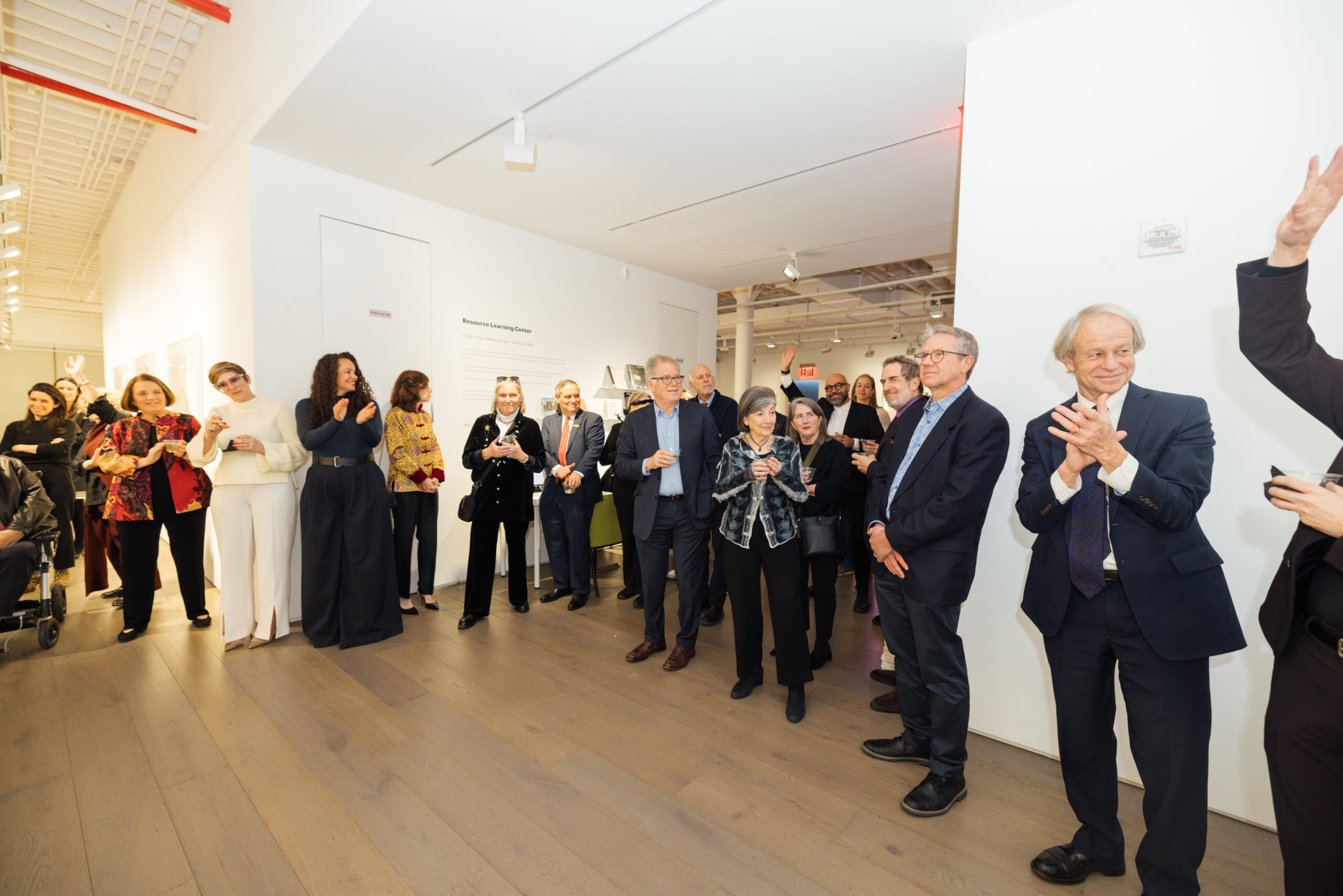 A group of people is gathered in a brightly lit event space, engaging in conversation and socializing. Some individuals are clapping or holding drinks, while others are looking towards a speaker off-camera. The atmosphere appears festive, with a mix of formal and casual attire, and a wall displaying information about a learning center in the background.
