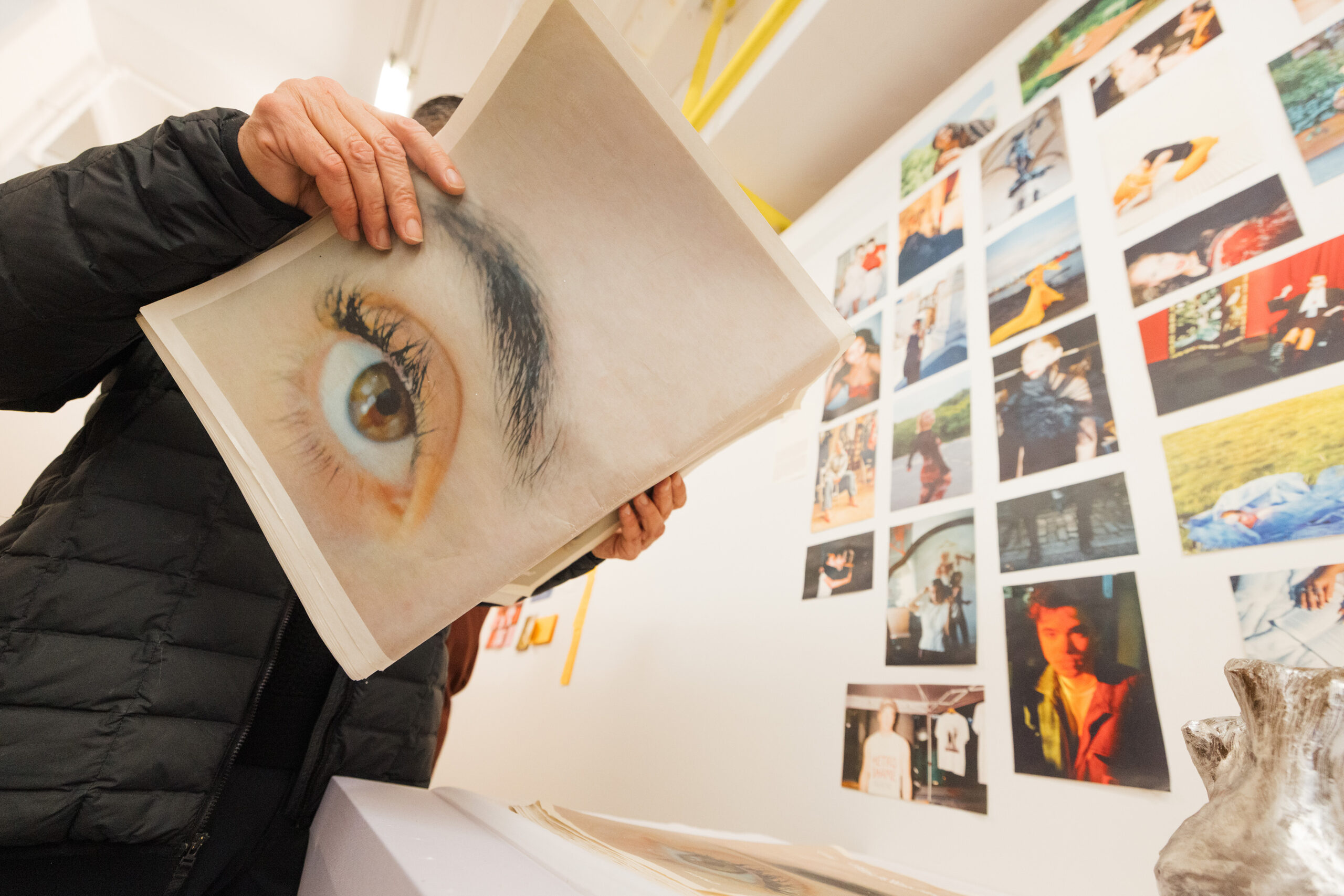 A person holds a large magazine page featuring a close-up of an eye, partially obscuring their face. In the background, a wall displays a collage of various photographs. The setting appears to be an art exhibit, with soft lighting and a contemporary aesthetic.