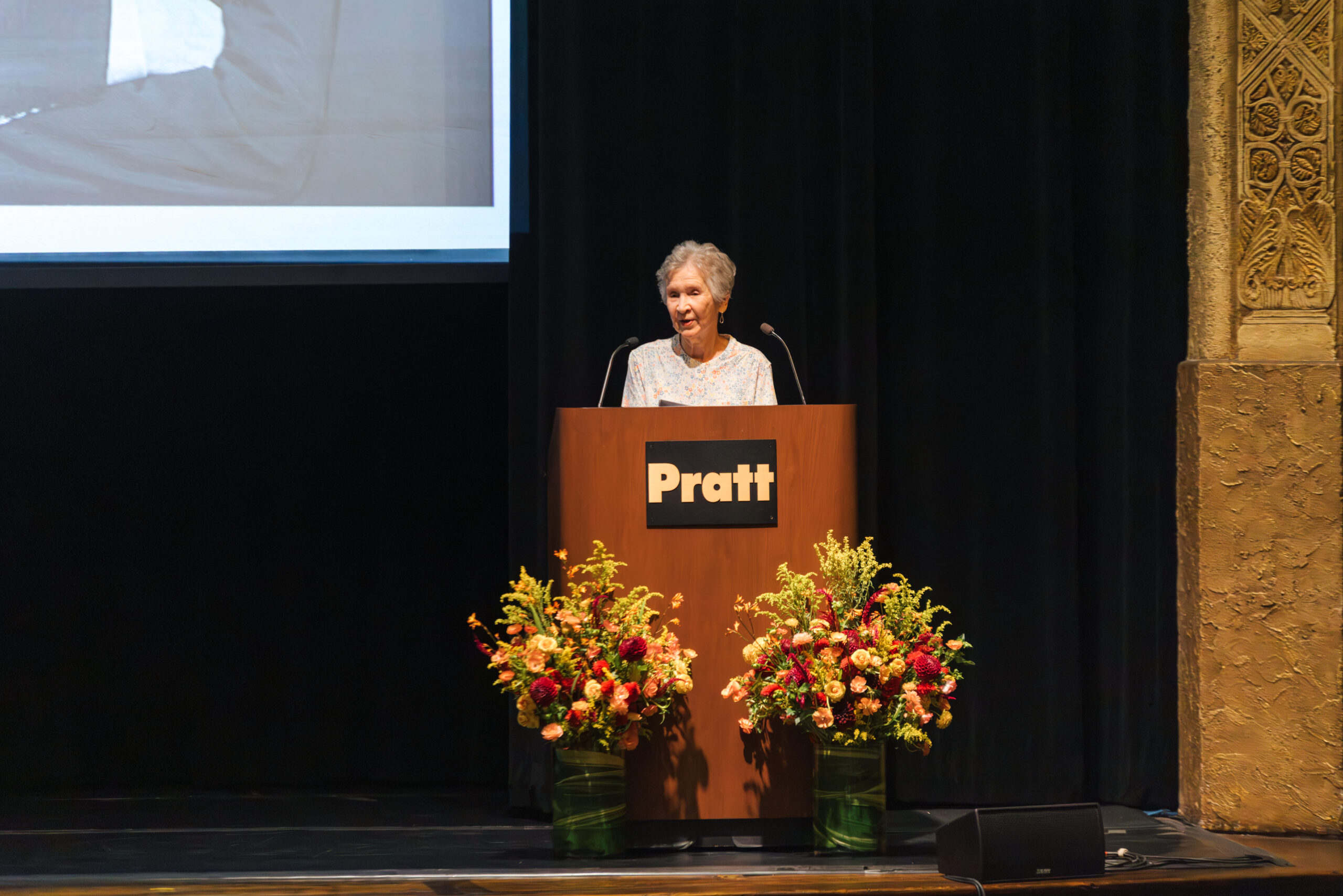 A woman with short, gray hair stands at a podium with a sign that reads "Pratt." She is speaking to an audience and appears to be presenting or giving a speech. The podium is adorned with two large floral arrangements in vibrant colors. The background is dark, highlighting the podium and speaker, with a large screen visible behind her displaying a black-and-white image.