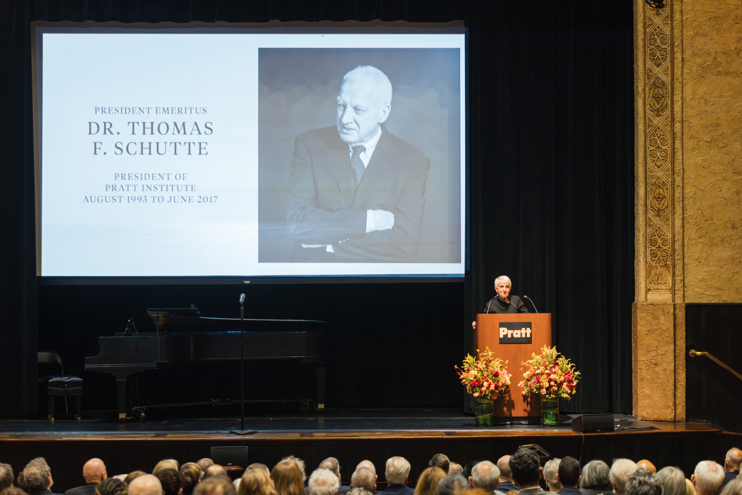 A speaker stands at a podium decorated with flowers, addressing an audience in a formal setting. Behind them, a large screen displays a portrait of Dr. Thomas F. Schutte, alongside text identifying him as President Emeritus of Pratt Institute from August 1993 to June 2017. A grand piano is partially visible on the stage, and the audience is composed of individuals with diverse hair colors and styles, attentively listening.