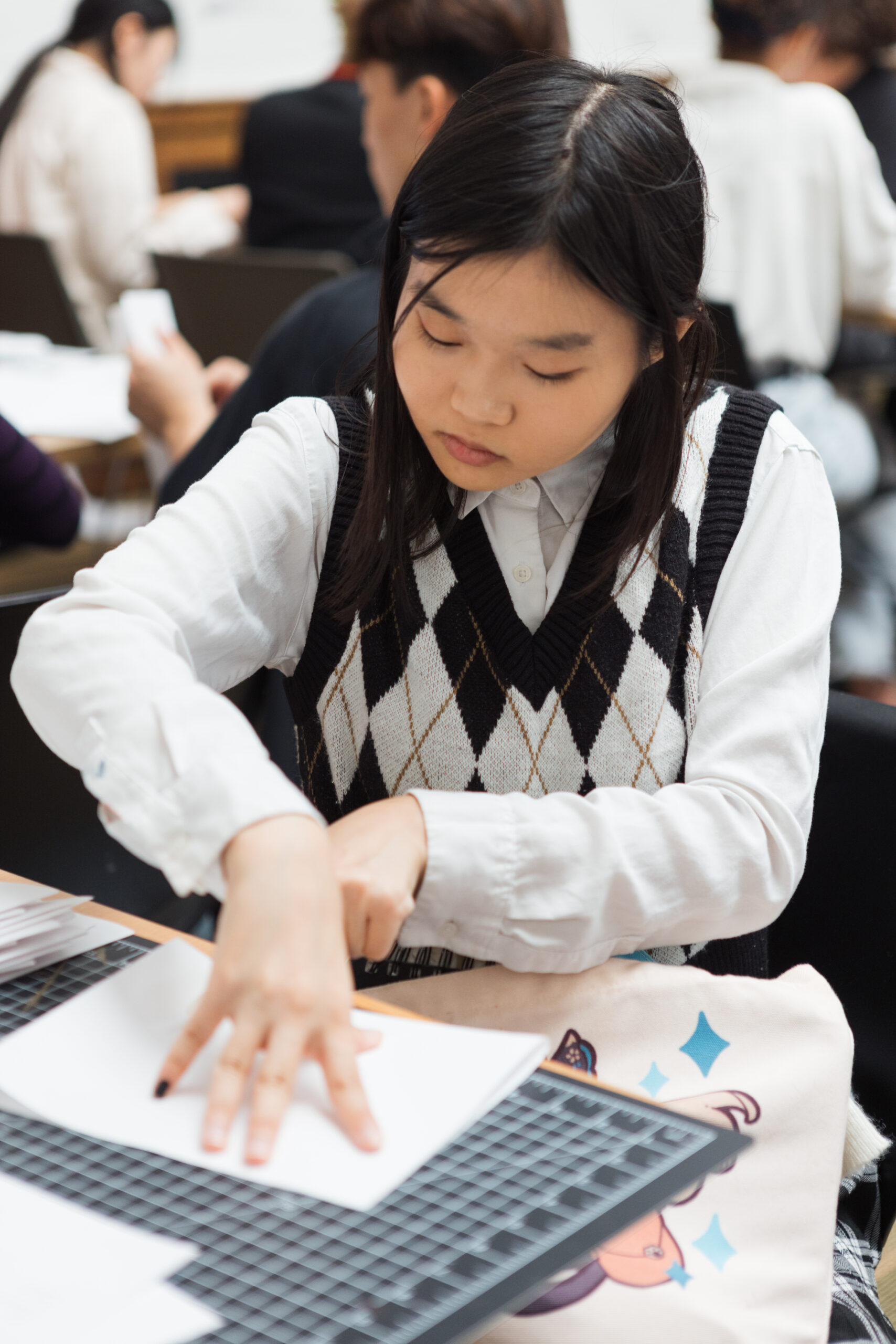 A young woman with long, dark hair is focused on cutting paper at a table. She wears a white shirt under a black vest with a diamond pattern. Her left hand holds down the paper while her right hand uses scissors. In the background, other individuals are engaged in similar activities, suggesting a creative workshop environment.
