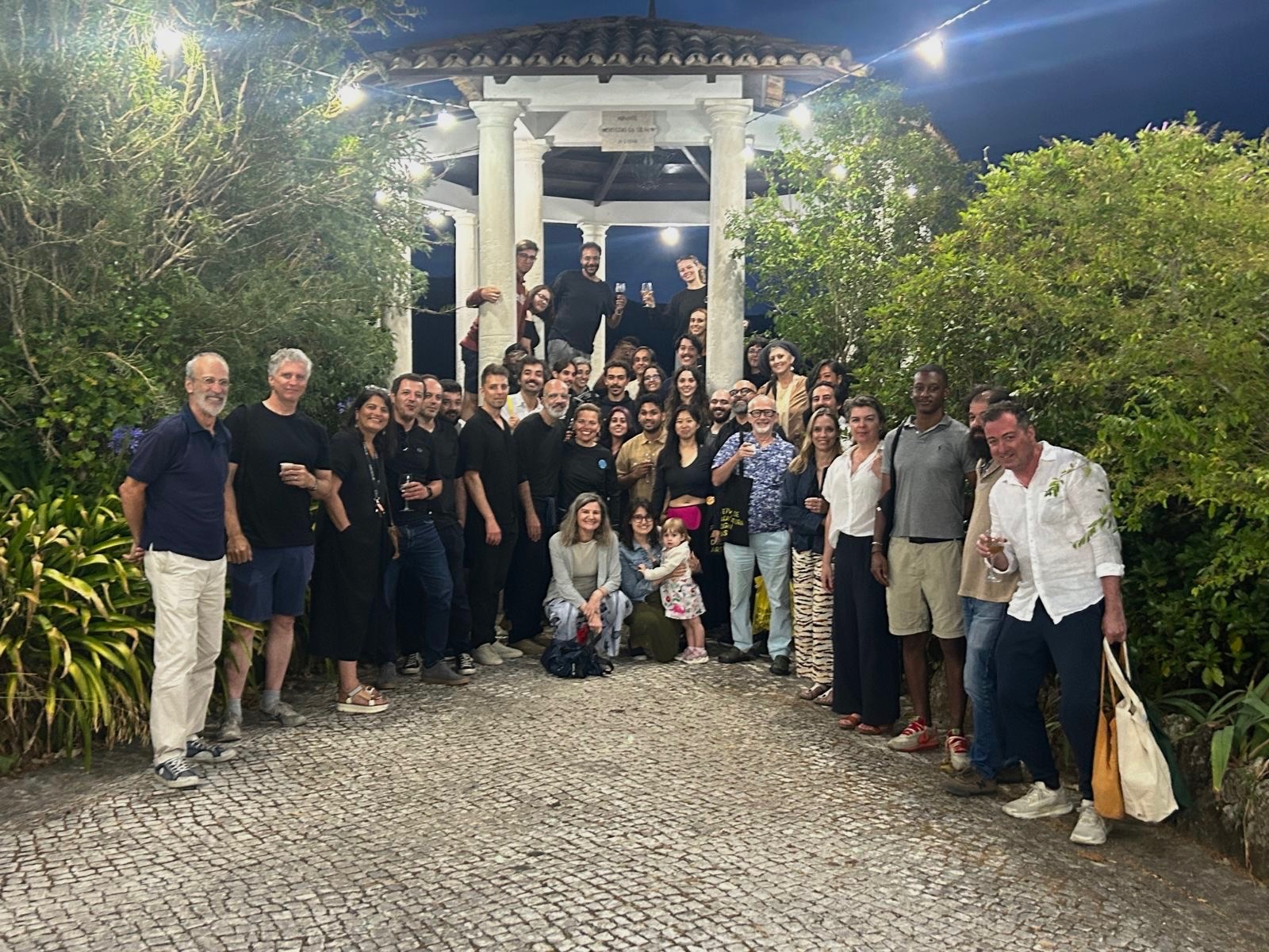 A large group of people stands in a garden setting, with a gazebo in the background. The individuals are diverse in appearance and attire, some smiling and posing for the camera. Greenery and decorative lights enhance the festive atmosphere. The ground is cobblestone. A few people are seated in front, while others are arranged standing around them, showcasing a casual gathering or celebration.