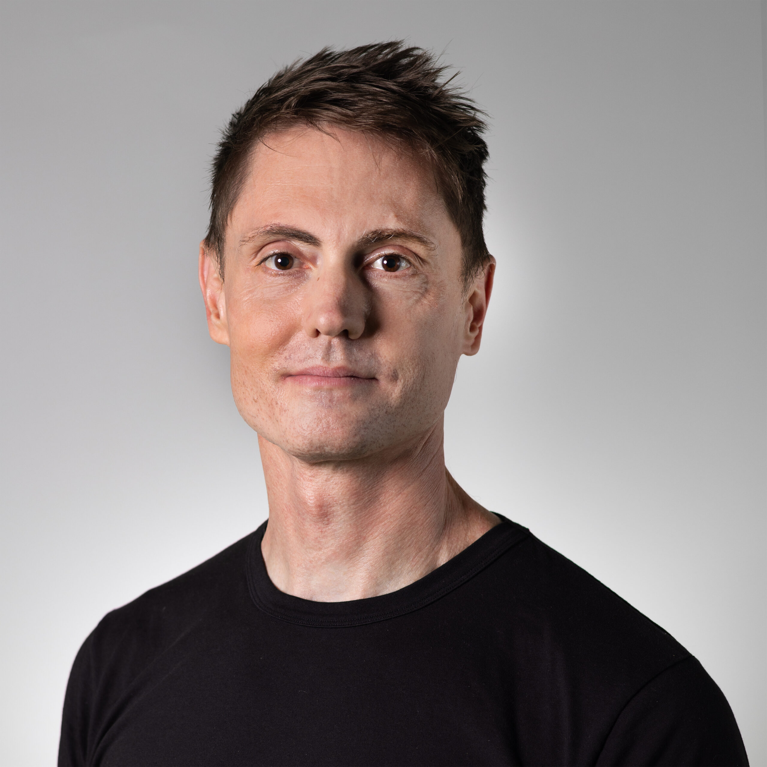 A close-up portrait of a person with short, tousled hair, wearing a black T-shirt. They have a neutral expression and appear to be facing directly towards the camera. The background is a smooth gradient from light to dark gray.