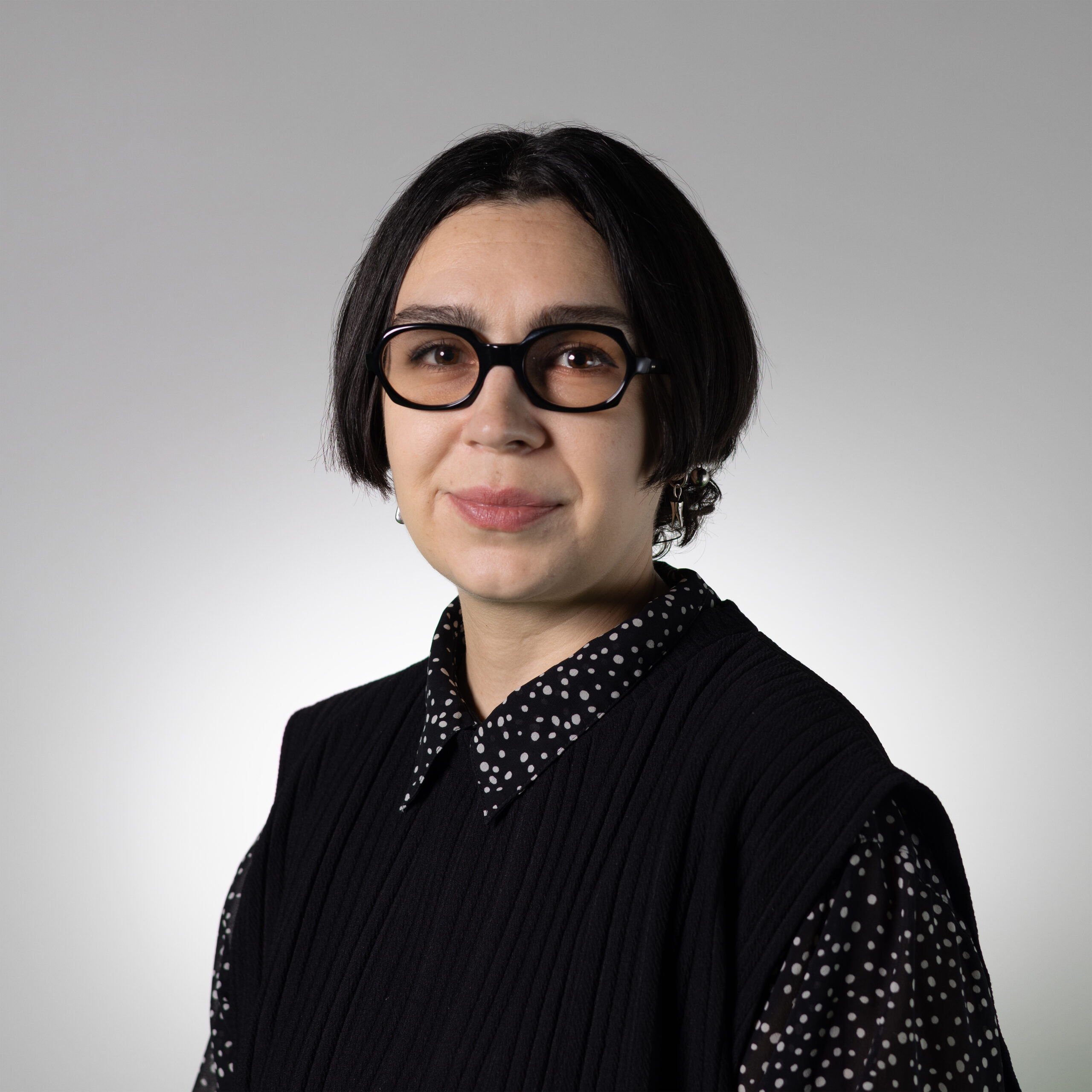 A person with short, dark hair and glasses smiles at the camera. They are wearing a black textured top over a collared, polka-dotted shirt. The background is a light gradient from white to gray, creating a soft, neutral setting.