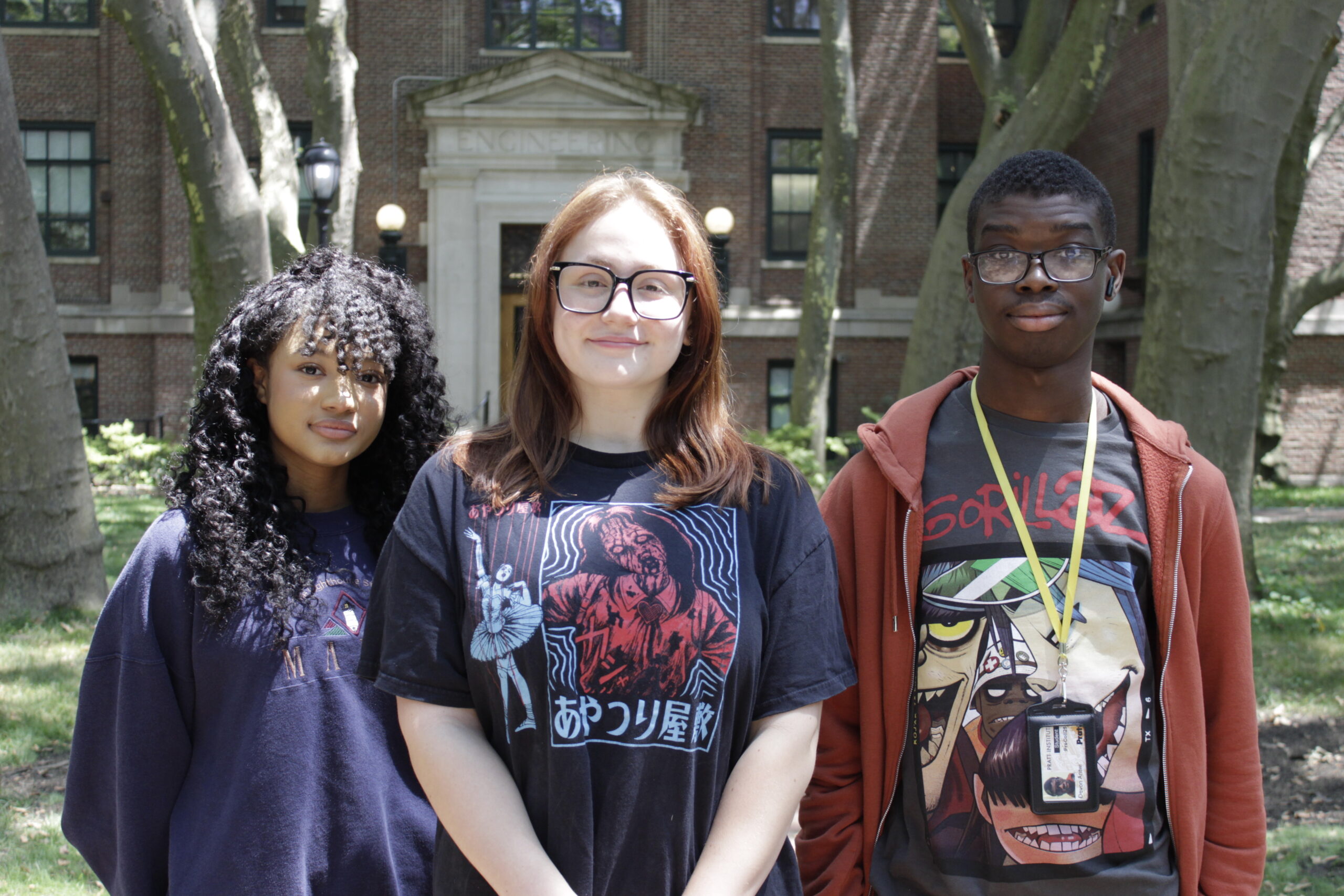 Three young adults stand together outdoors in front of a brick building with the word "Engineering" above the entrance. The woman in the middle wears glasses and a graphic black t-shirt featuring a design with a figure. The person to her left has curly hair and wears a dark sweater, while the person on her right wears a red hoodie over a black t-shirt with colorful artwork. Sunlight filters through trees in the background.