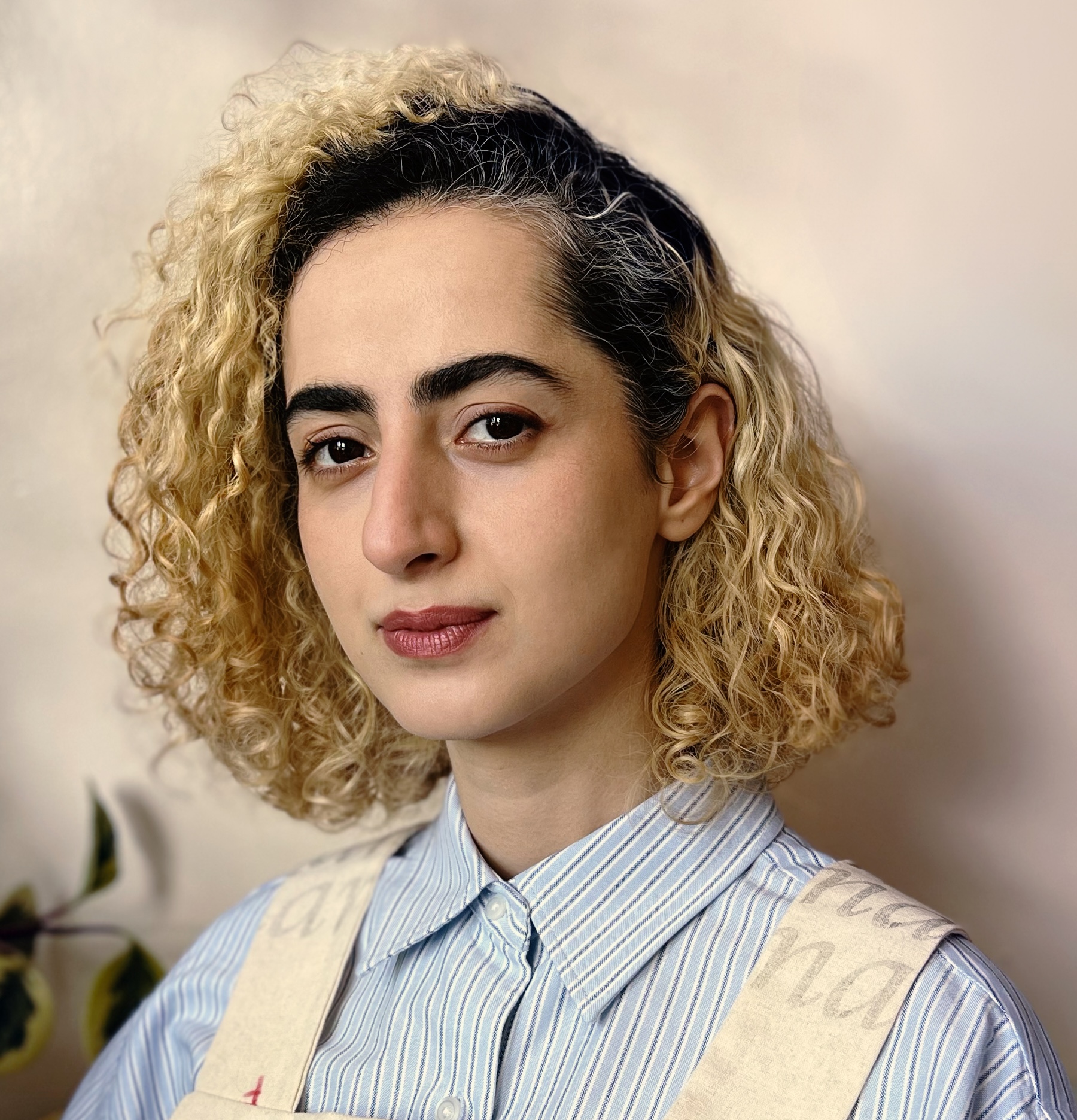 A close-up portrait of a young person with curly, blonde hair and a neutral expression. They are wearing a light blue striped shirt and an apron with text on it. The background is softly blurred with hints of beige and green, suggesting a warm ambiance.