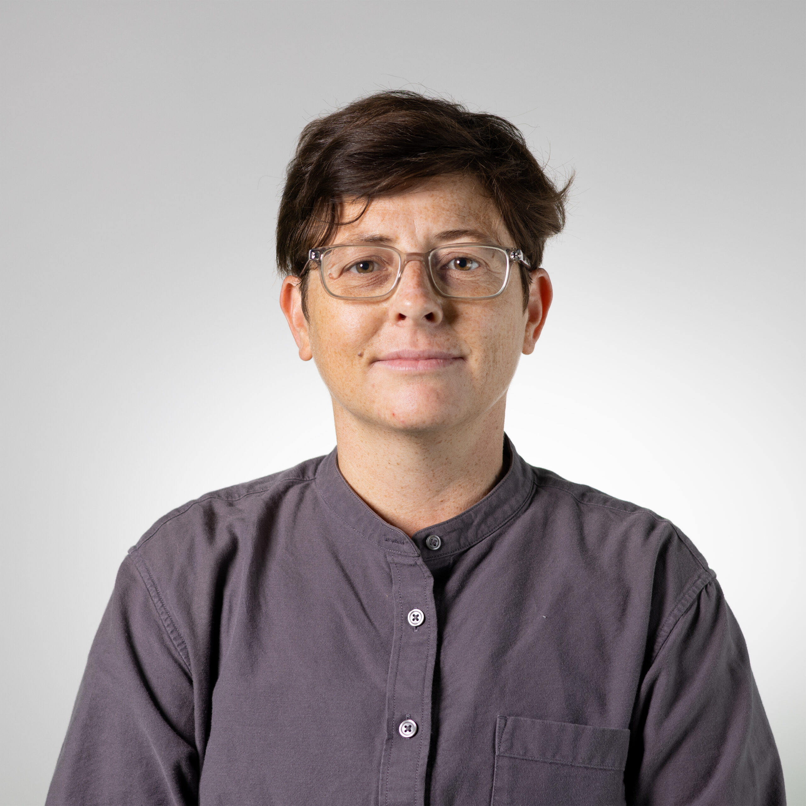 A person with short, tousled brown hair and glasses is facing the camera, wearing a dark gray collared shirt. The background is a soft gradient from light gray to white. The person has a neutral expression.