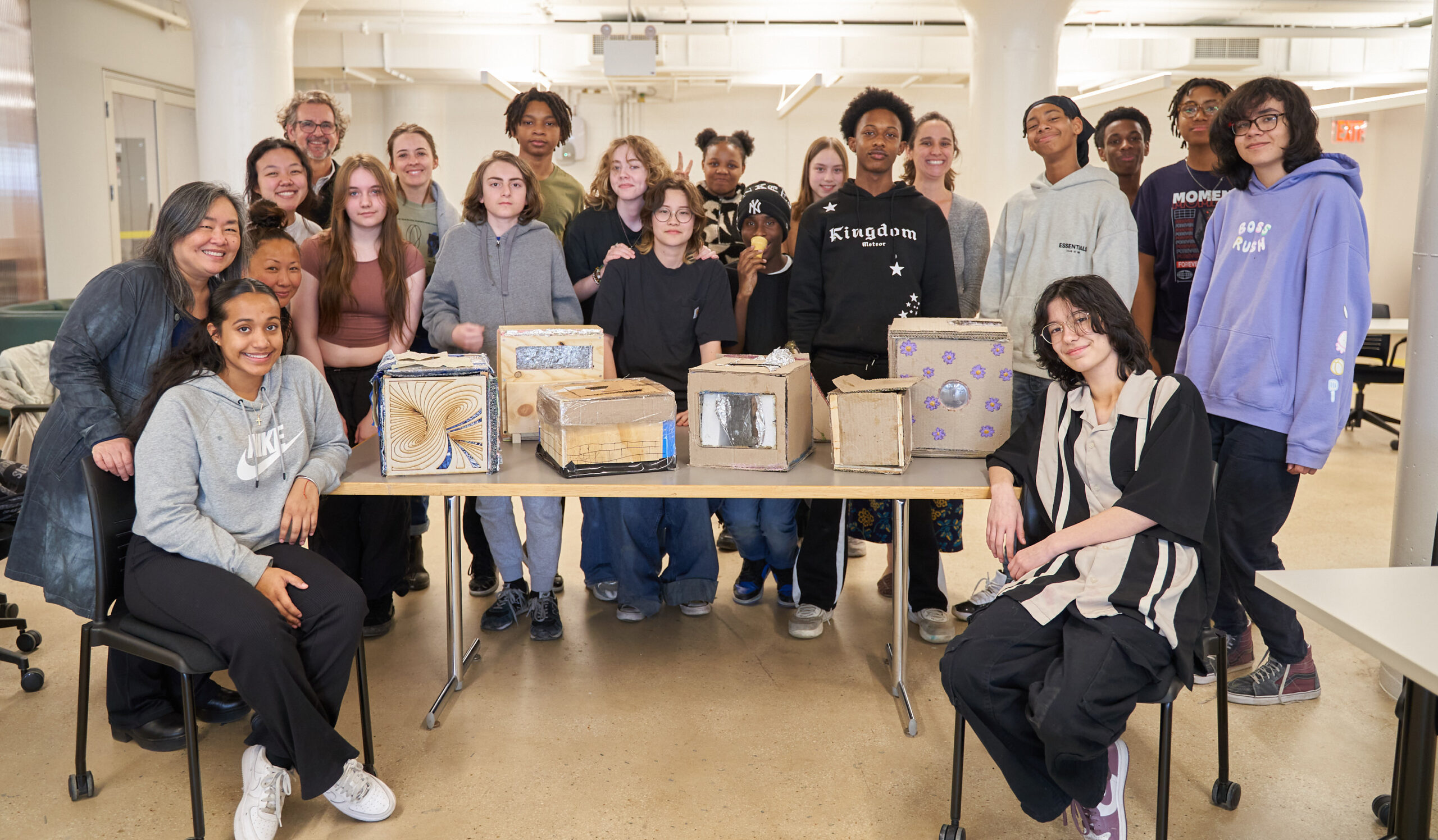 A diverse group of students and adults pose together in a bright indoor space, standing and sitting around a table. The table features several creatively decorated boxes made from cardboard. The group appears cheerful and engaged, with some members smiling directly at the camera. The setting includes large windows, bright lighting, and a modern design.