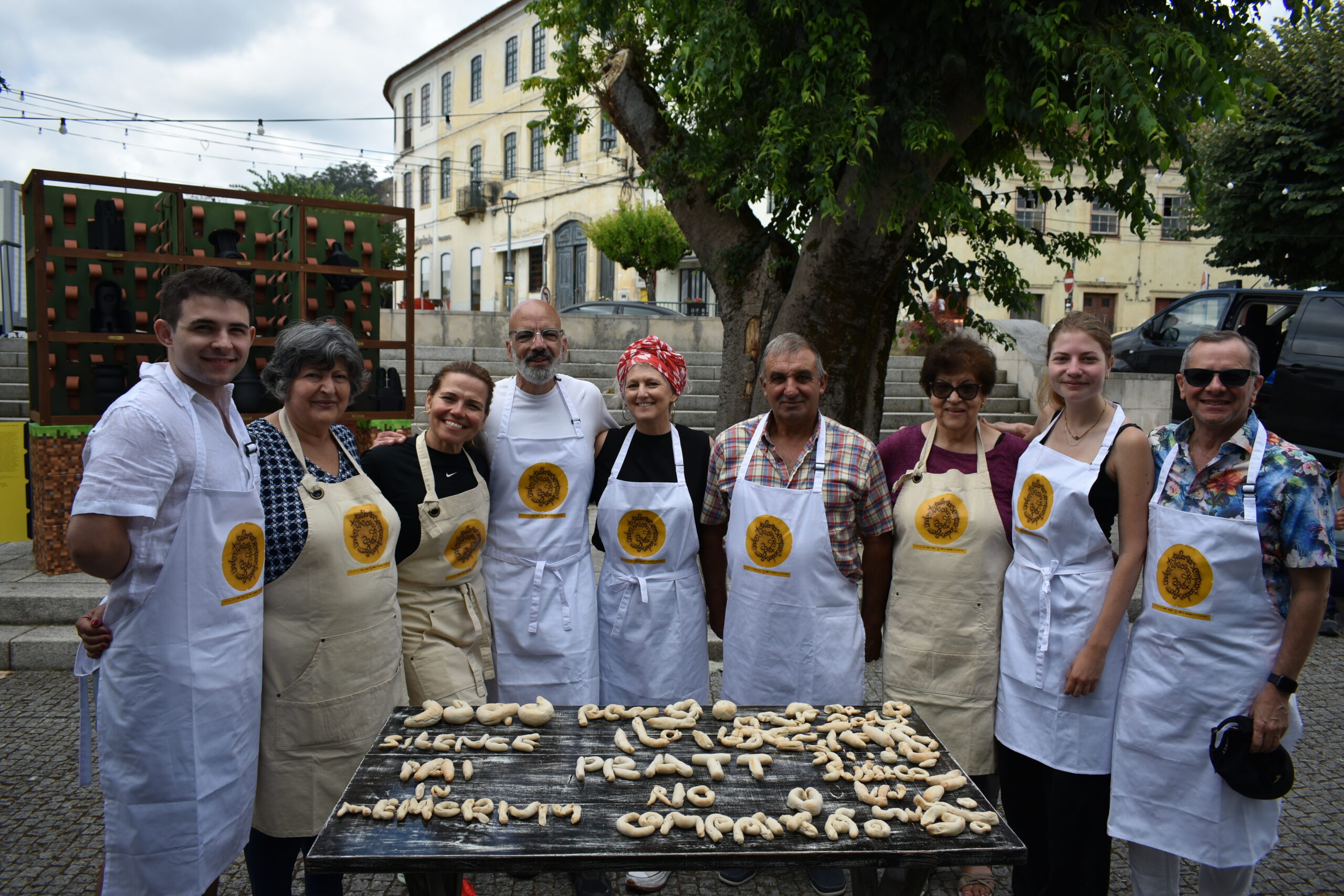 A group of nine individuals poses together outdoors, all wearing aprons with a circular logo. They stand behind a wooden table covered with dough shaped into various forms. In the background, there are stone steps, greenery, and a historical building. The atmosphere appears casual and inviting, suggesting a cooking or baking activity.