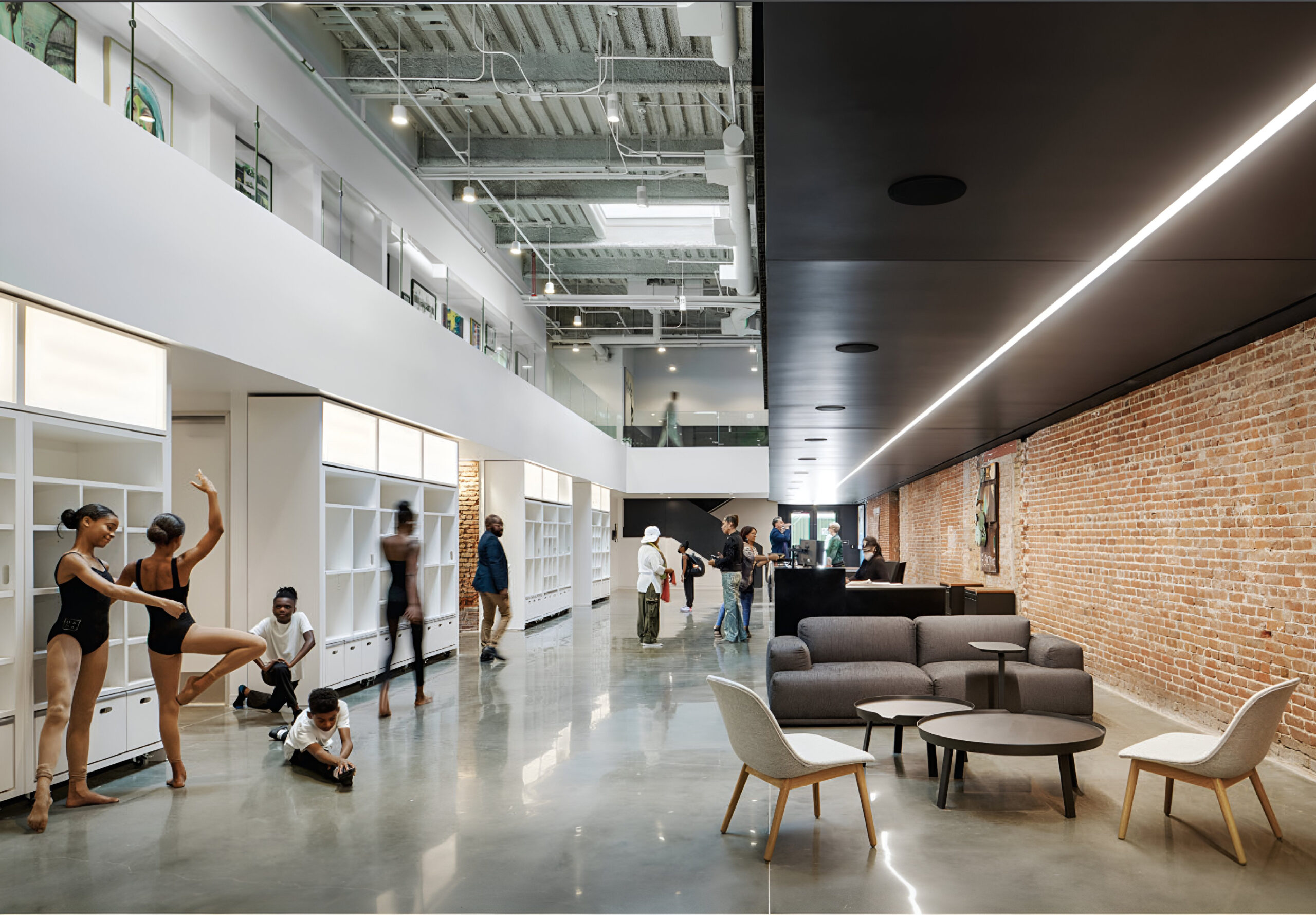 A spacious modern lobby featuring a mix of dance students and adults engaged in various activities. Two dancers in black leotards practice poses while another child plays on the floor. Several people are gathered near a reception area, with a brown brick wall and large windows in the background. The space is well-lit with a sleek design, showcasing comfortable seating arrangements.