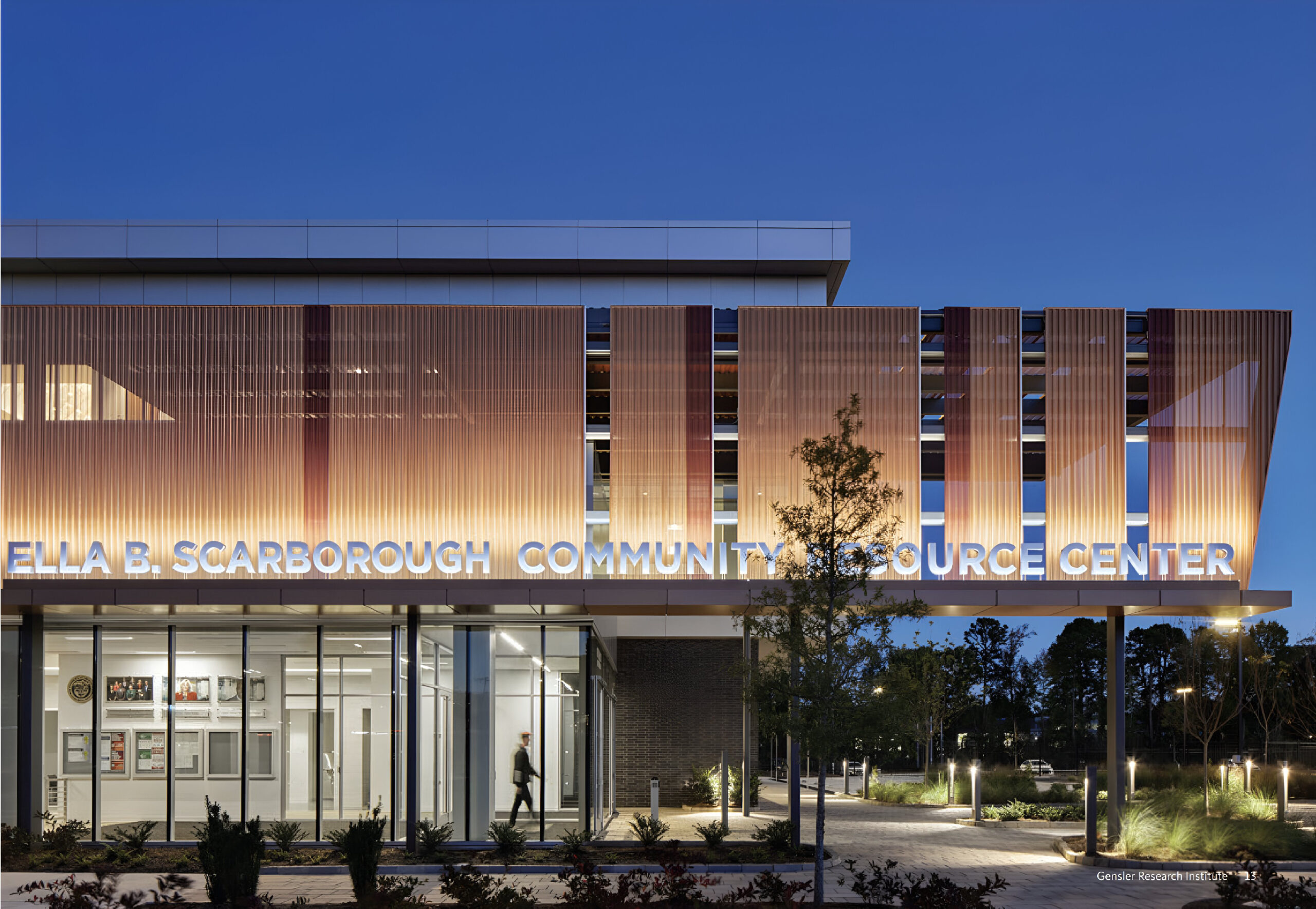 The image shows the exterior of the Ella B. Scarborough Community Resource Center at dusk. The building features modern architecture with large glass windows and a facade adorned with vertical slats, illuminated by soft lighting. The name of the center is prominently displayed in white lettering at the front, while a person walks by in the foreground. Surrounding greenery and pathway lighting enhance the inviting atmosphere.