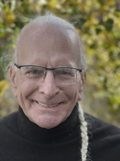 Head-and-shoulders color portrait of an older adult with light skin, wearing glasses and a black sweater, smiling gently while standing outdoors. The background is softly blurred with green and yellow foliage, suggesting a natural setting in daylight.