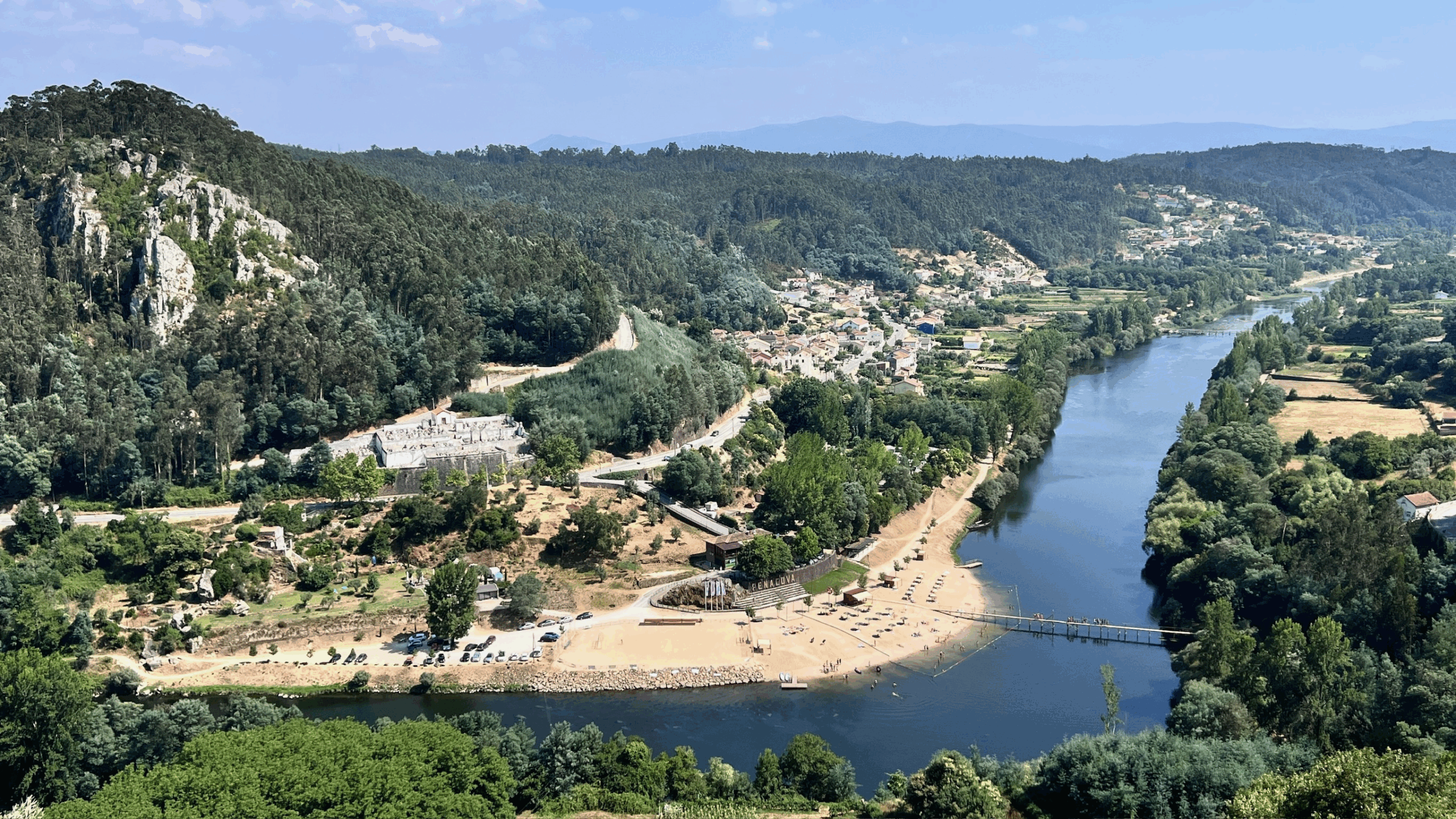 A panoramic view of a lush landscape featuring a river winding through greenery. On the left, a rocky hill is covered with trees. In the foreground, a sandy area with beach umbrellas and chairs borders the river. To the right, several small houses and agricultural fields are visible against the backdrop of distant mountains under a blue sky.