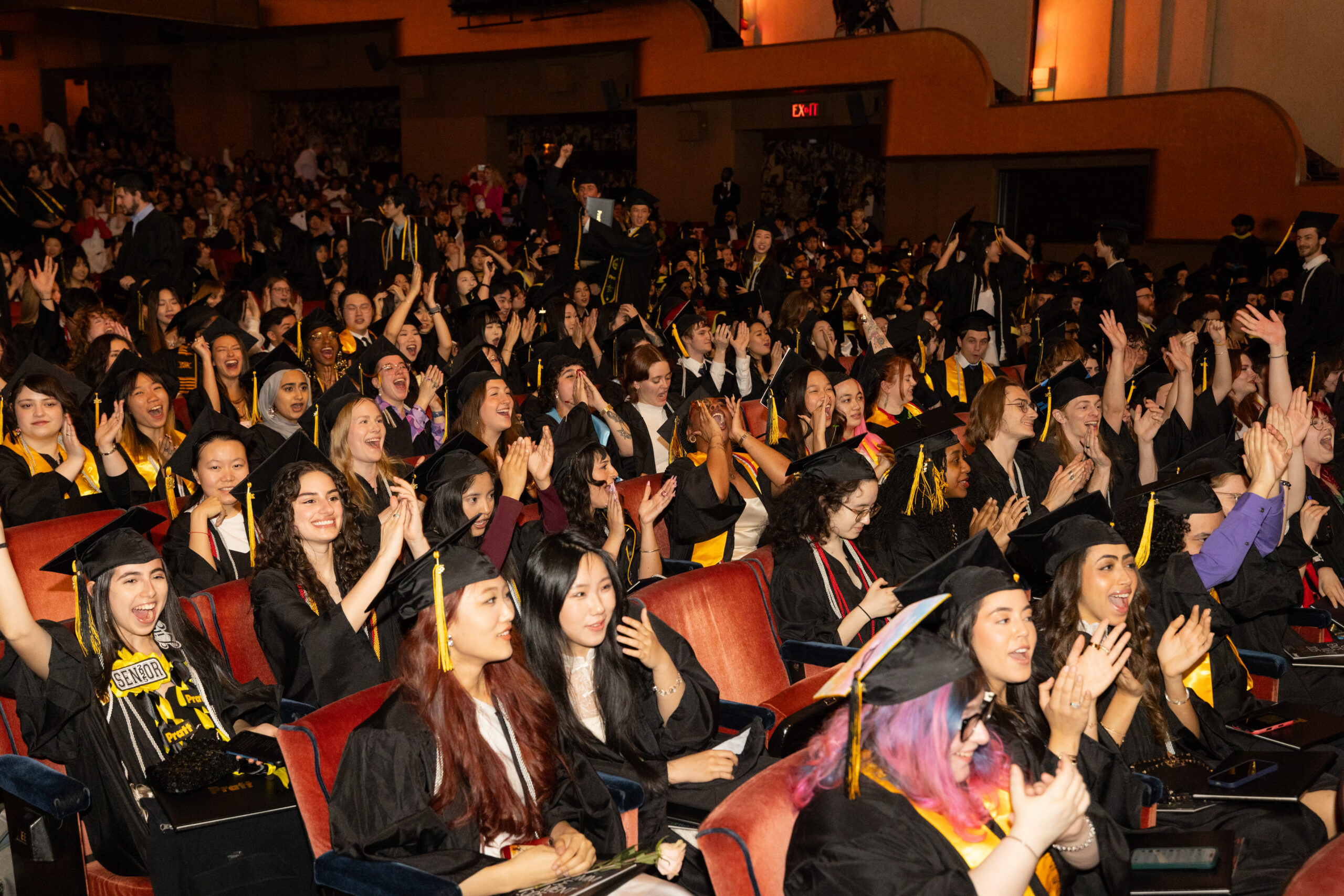 A large group of graduates in black caps and gowns is seated in an auditorium, enthusiastically clapping and celebrating. Many are smiling and cheering, creating a lively atmosphere. The crowd includes a diverse mix of individuals, with some holding signs or flowers. The venue has an elegant design, with warm lighting and a backdrop of cheering friends and family.