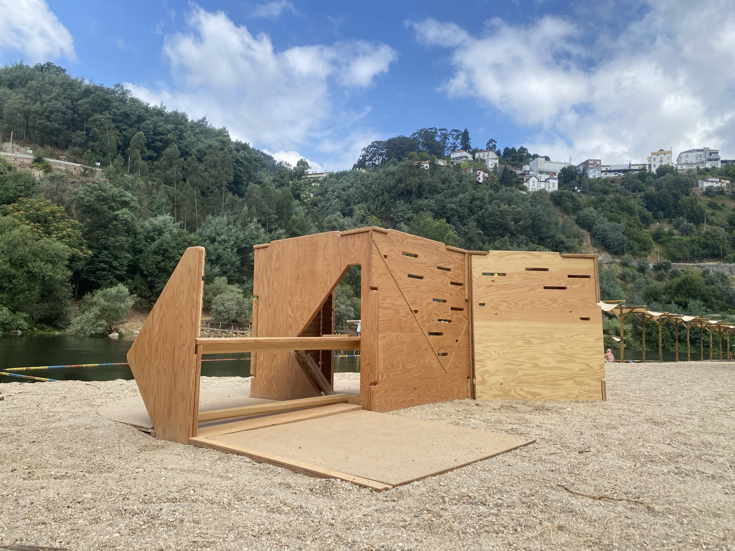 A wooden structure resembling a small pavilion or shelter is positioned on sandy ground near a river. The exterior features angled walls with cut-out shapes and a flat roof, surrounded by greenery and hills in the background. Scattered trees and some buildings are visible on the hillside, with a few people in the distant area. The sky is partly cloudy.