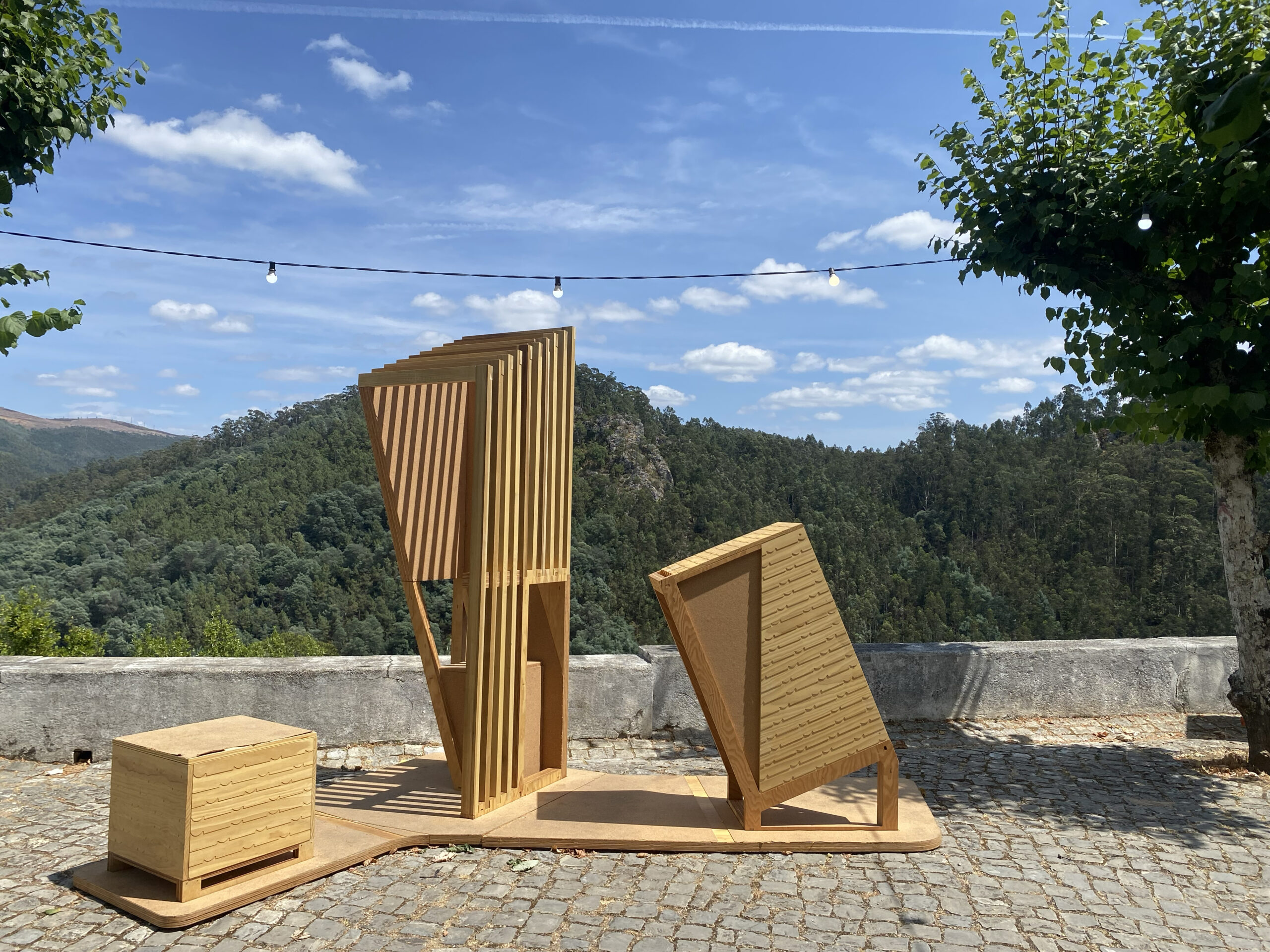 An artistic outdoor furniture installation made of wood, featuring a tall, angular structure with vertical slats and an attached flat surface. Beside it, a small square table with textured drawers rests on a stone pavement. In the background, dense green hills and a clear blue sky with scattered clouds are visible. Decorative string lights are hanging above, and nearby trees provide shade.