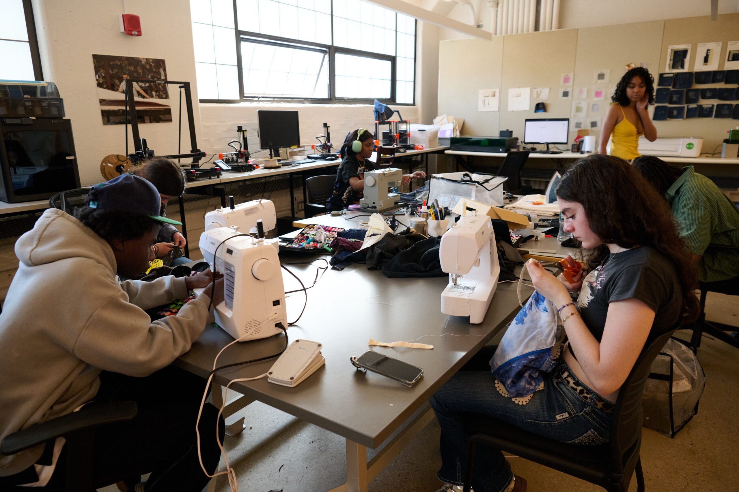 A group of students are seated around a large table in a bright classroom, working with sewing machines and fabric. Some students are focused on hand stitching, while others are using the machines. Spools of thread, fabric pieces, and sewing supplies are spread across the table. In the background, computers, 3D printers, and design materials suggest a creative lab environment. A student in a yellow dress stands near the back wall observing the activity.