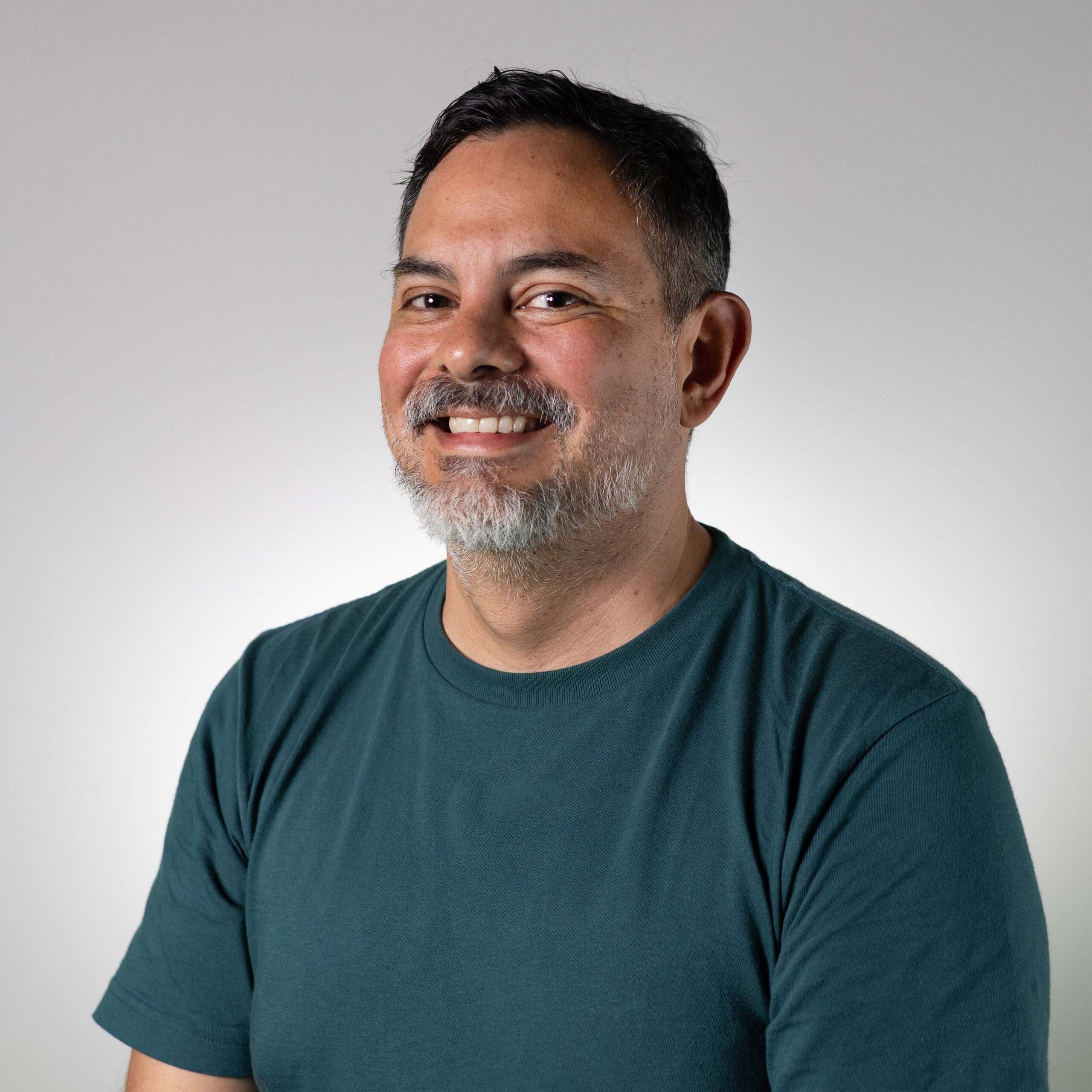 A person with short, dark hair and a salt-and-pepper beard smiles while wearing a dark green t-shirt. The background is a soft gradient from white to light gray.
