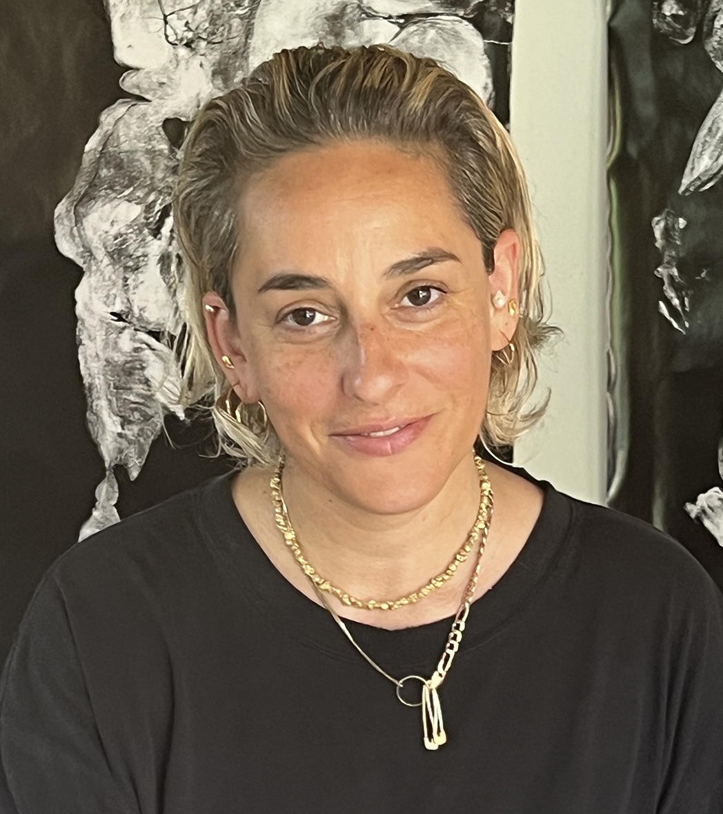 A person with short, wavy blonde hair is smiling at the camera. She wears a black shirt and multiple gold necklaces. The background features abstract art in black and white.