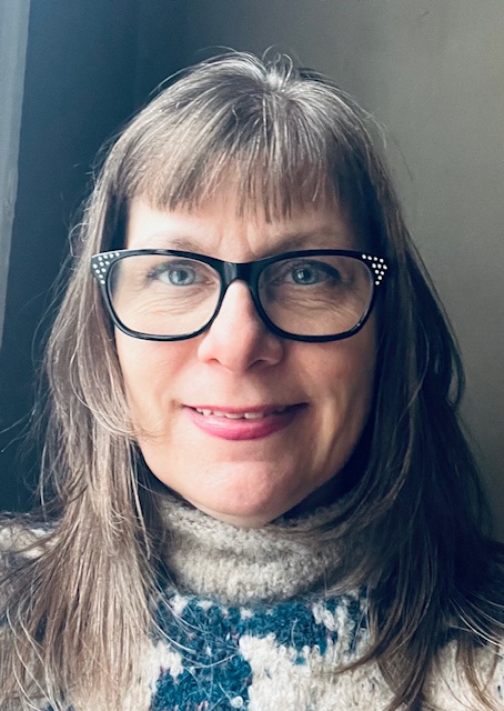 Head-and-shoulders portrait of a person with light skin wearing glasses and a knit sweater, photographed indoors near a window. The individual is smiling slightly, with soft natural light illuminating their face against a neutral background.