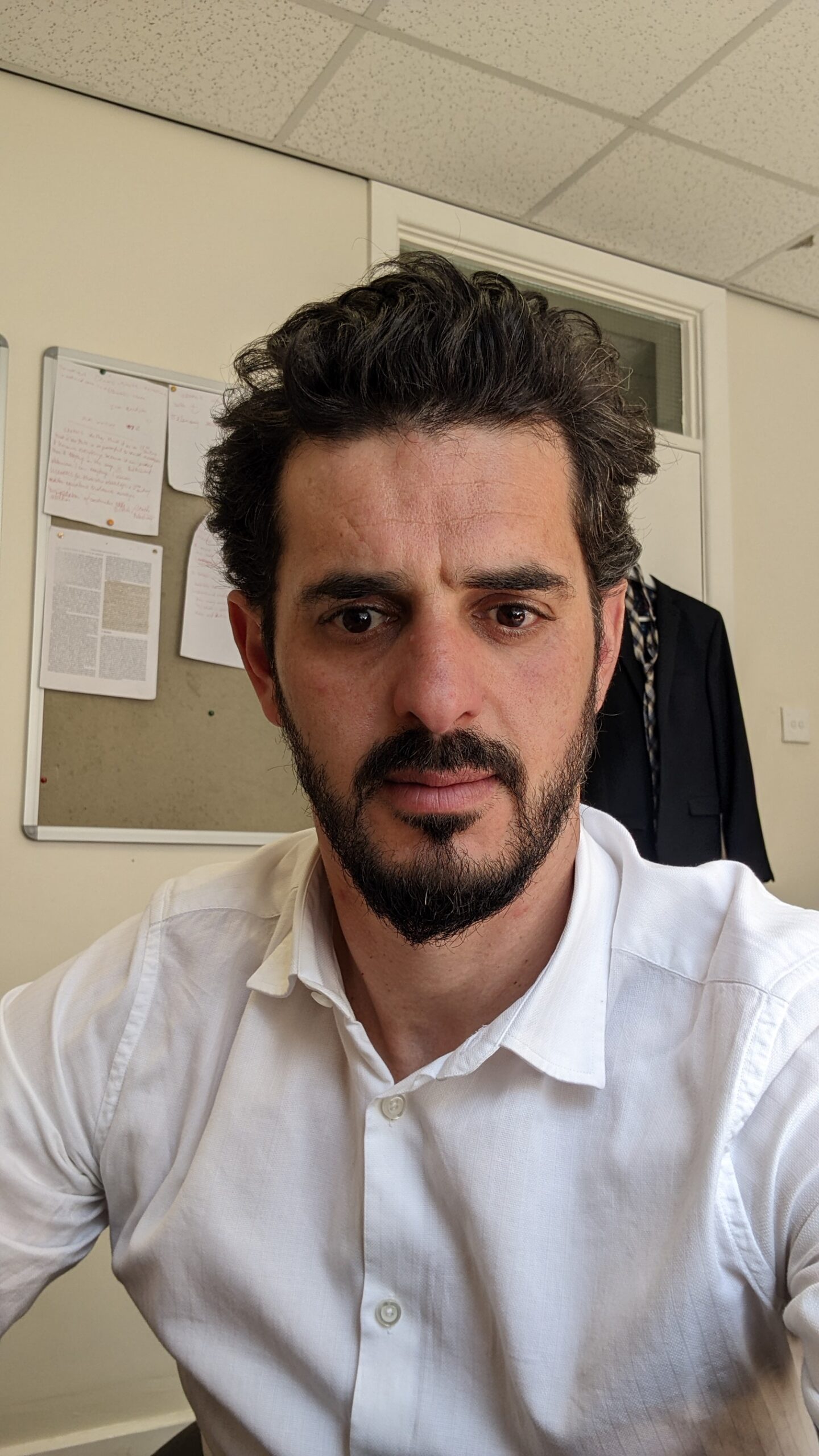 Close-up indoor portrait of a person with medium skin tone, dark hair, and a short beard, wearing a white button-down shirt. The individual faces the camera with a neutral expression. The background shows an office environment with a bulletin board, papers, and a hanging jacket.