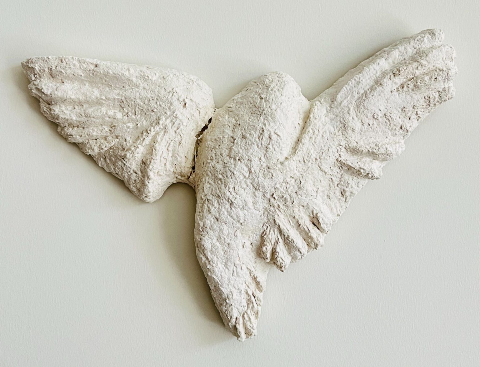 A textured white plaster sculpture of a bird in mid-flight with wings spread. The rough surface emphasizes the feather details, and the figure is mounted against a plain wall.