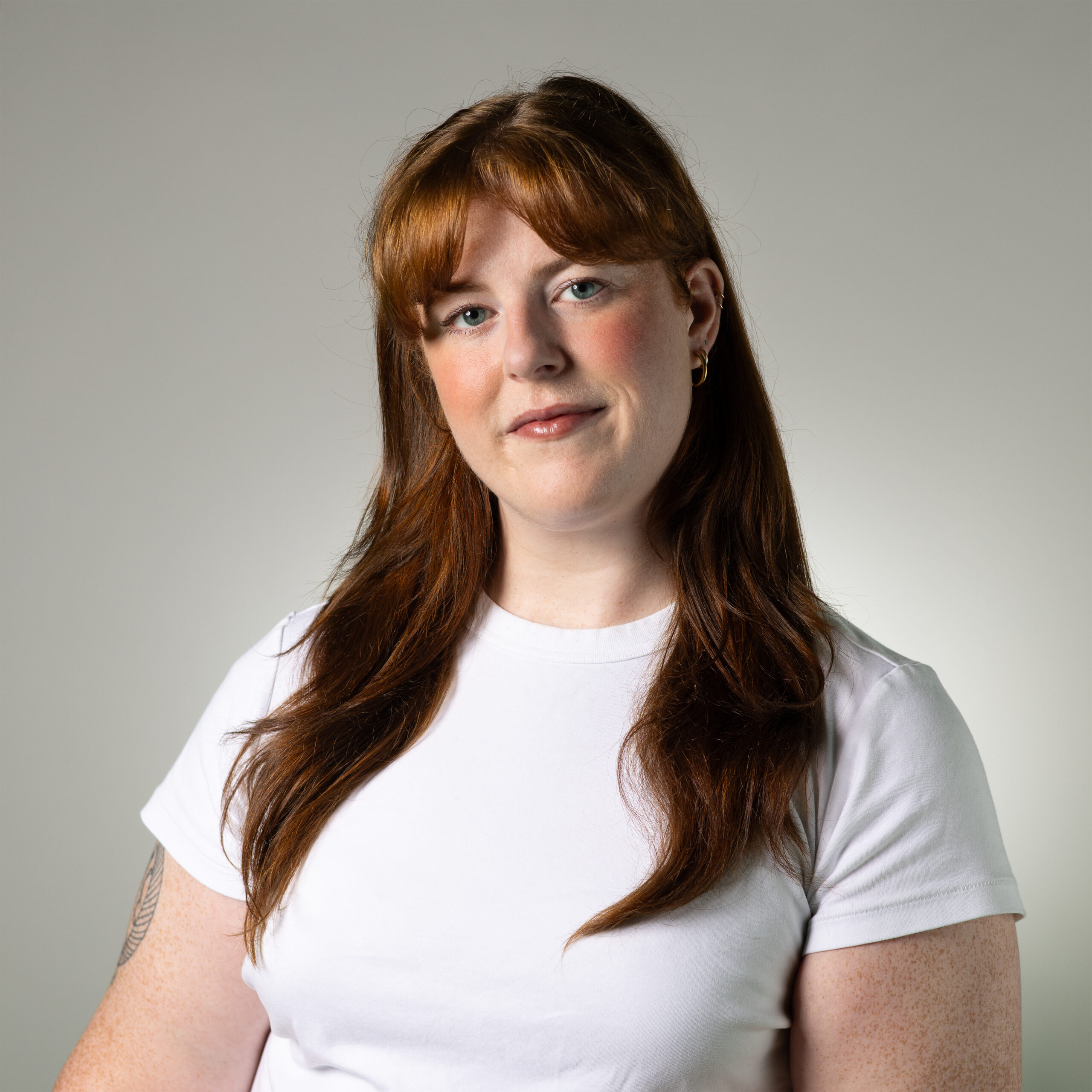 A young adult with long, wavy red hair stands against a plain gray background. They wear a simple white t-shirt and have minimal makeup, smiling gently. Freckles are visible on their arms. The lighting is soft and highlights their features.