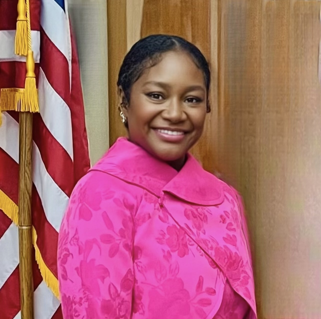 A person is smiling while standing next to a wooden surface and an American flag. They are wearing a bright pink floral coat with a high collar. Their hair is styled in a low bun, and she has earrings. The setting appears official, suggesting a professional or ceremonial context.