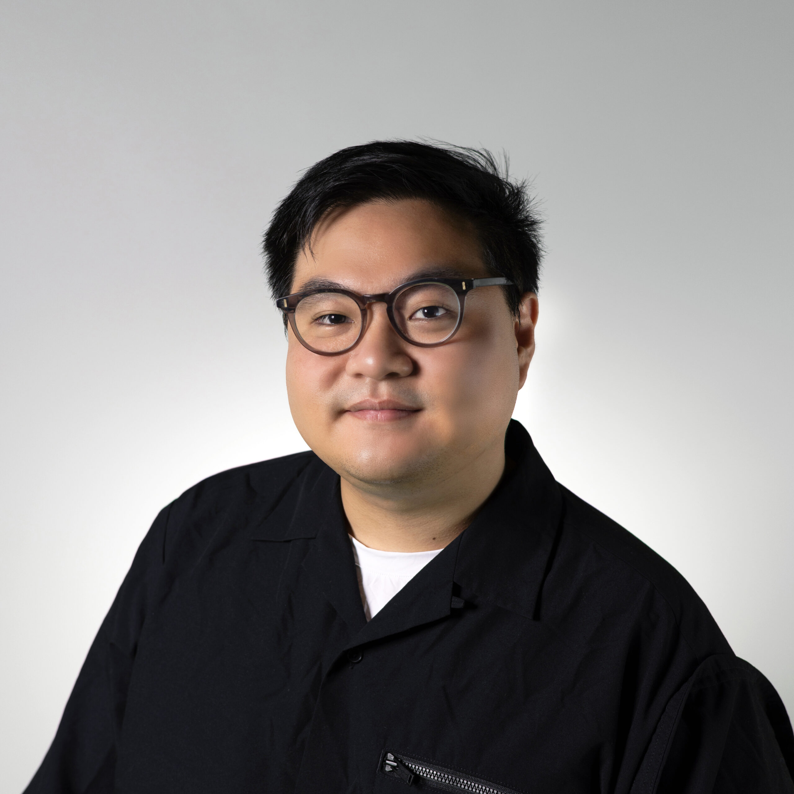 A person with short, dark hair and round glasses smiles slightly at the camera. They are wearing a black shirt with a zippered pocket. The background is a light gradient, transitioning from white to gray.