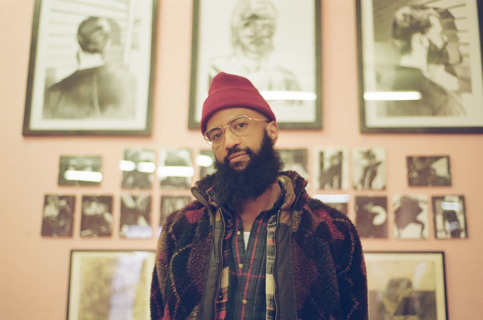 A man with a full beard and glasses stands indoors against a wall covered in framed drawings and photographs. He is wearing a red knit beanie, plaid shirt, and a patterned jacket. The background features large portrait sketches above smaller black-and-white photographs arranged in a grid. The overall setting appears to be an art gallery or exhibition space.