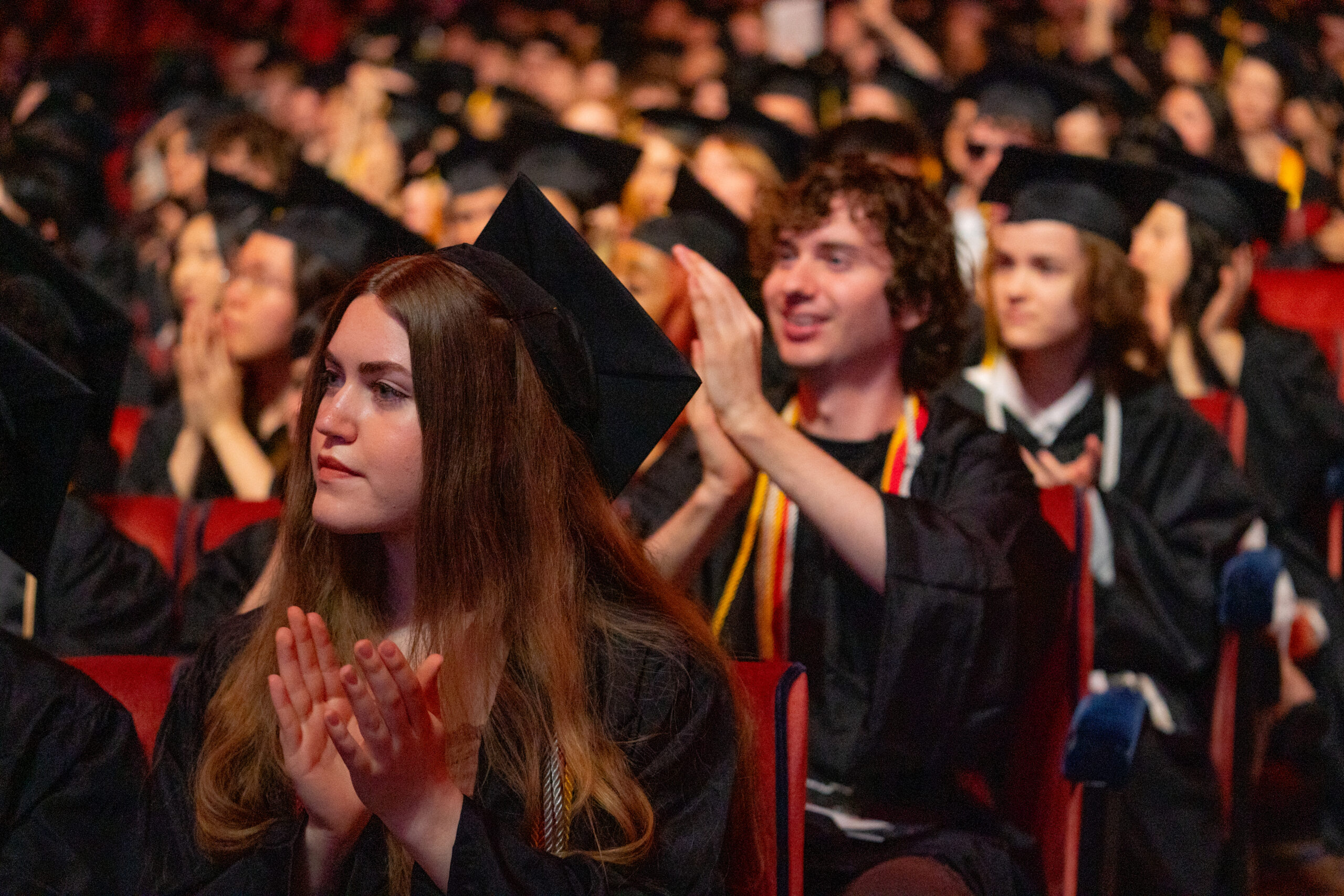 A crowded auditorium filled with graduating students wearing black caps and gowns. In the foreground, a woman claps with a focused expression, while a man behind her claps enthusiastically. The audience is engaged, with some students visible in the background, celebrating the graduation event.