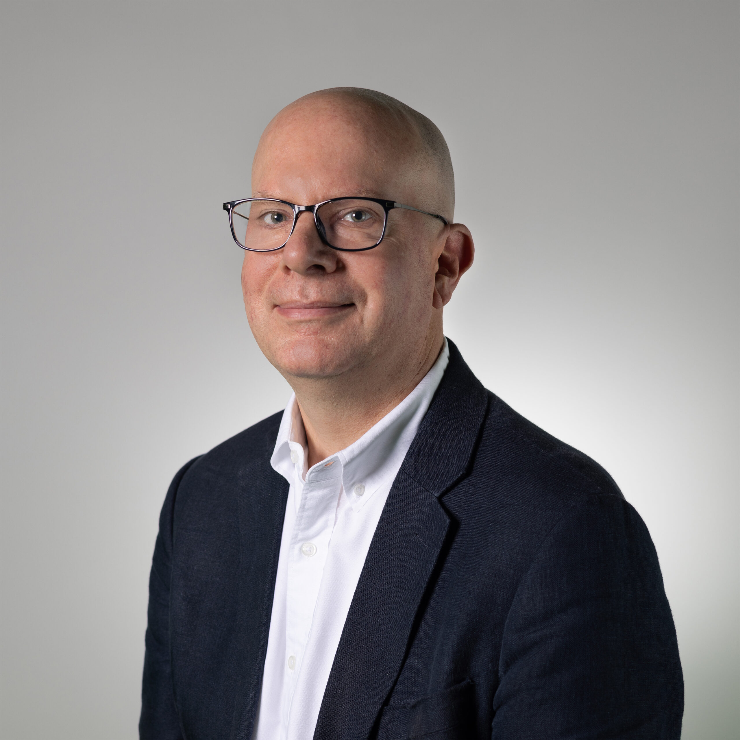 A professional-looking individual with a bald head and glasses is smiling slightly. He is wearing a dark blazer over a white collared shirt. The background is a light gray gradient, creating a clean and modern appearance.