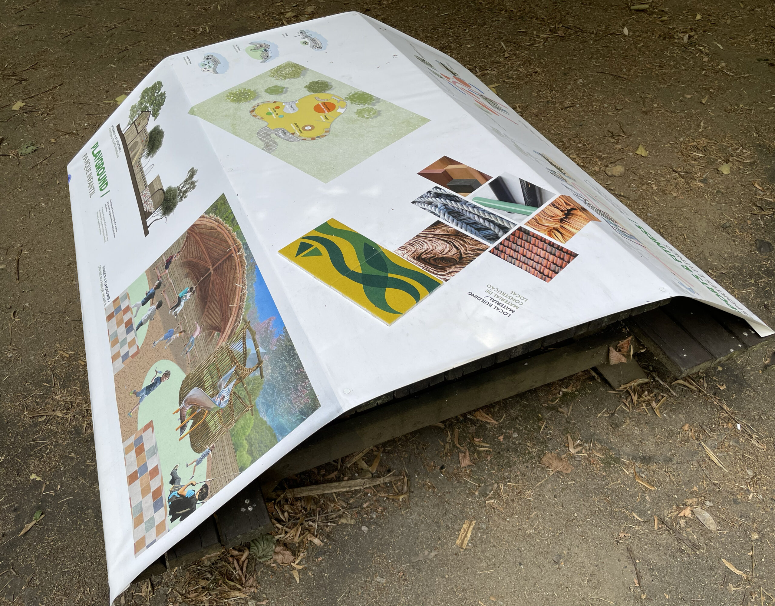 A shaded outdoor area features several wooden picnic tables, each with informational posters laid upon them. The posters include the word "VERDEVIVO" prominently displayed. In the background, there is a small brick structure that appears to be a barbecue or oven. A person is visible sitting at one of the tables, engaged with materials in front of them. The setting is surrounded by trees and dry foliage.