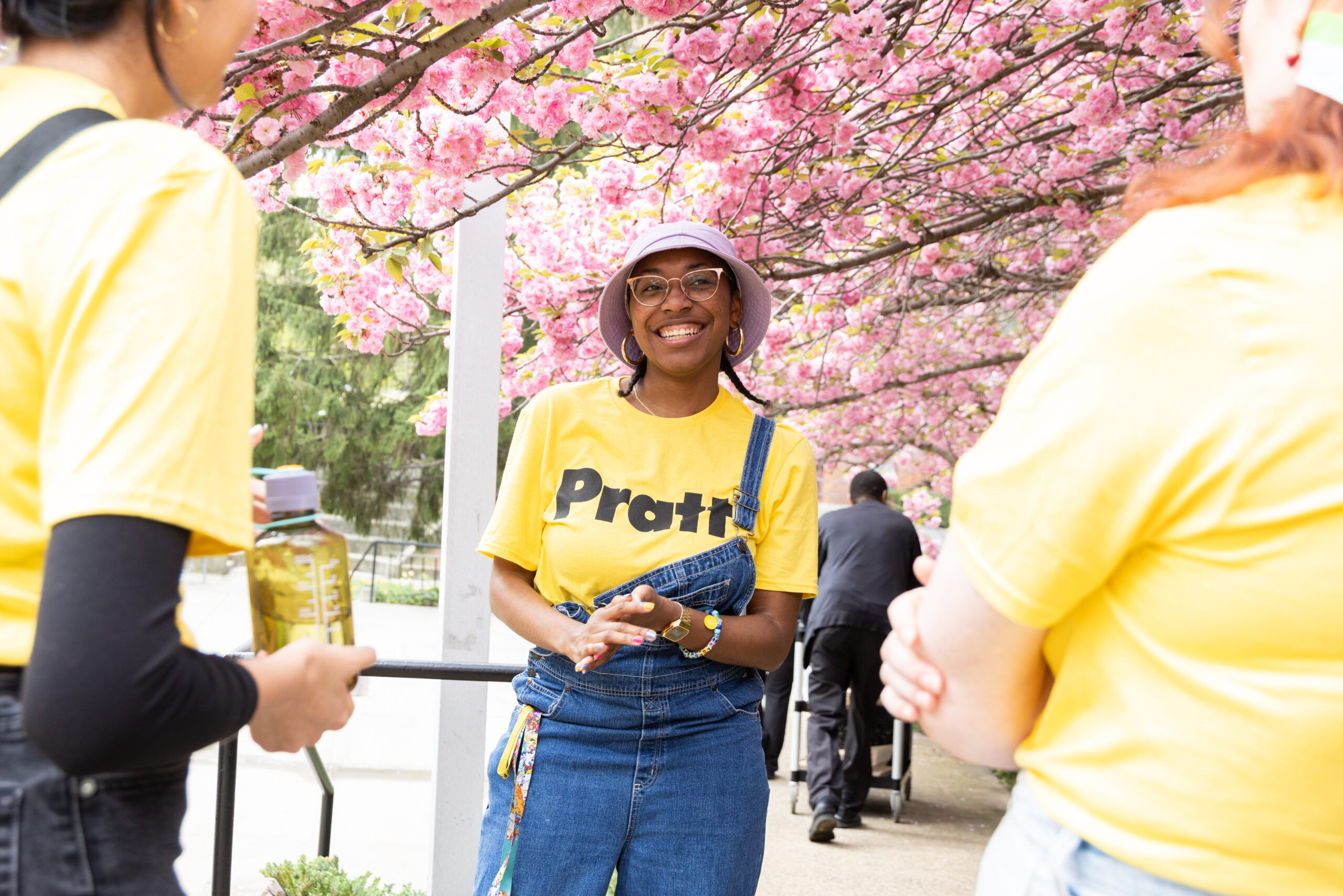 A smiling young woman stands in front of pink cherry blossom trees, wearing a yellow shirt with "Pratt" printed on it and overalls. She is engaging with two other people, one holding a water bottle. The scene captures a vibrant outdoor setting filled with flowers, suggesting a social event or gathering.
