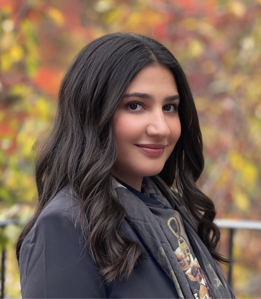 A person with long, wavy dark hair is smiling softly, wearing a dark blazer and a patterned scarf. They are outdoors, with a blurred background of autumn leaves in warm hues of orange, yellow, and green.
